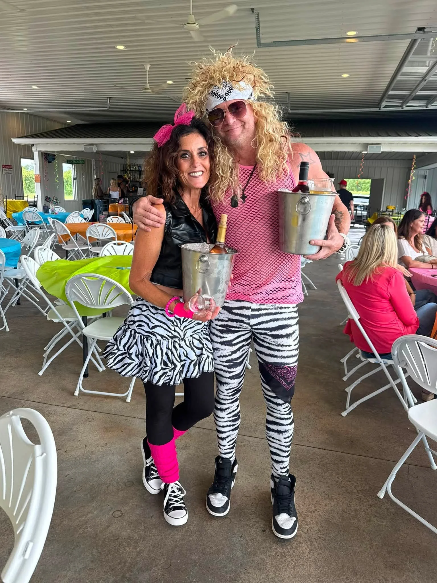 Two people in zebra print outfits posing with drinks in metal buckets at a party or event indoors with tables and chairs, some people sitting and chatting in the background.