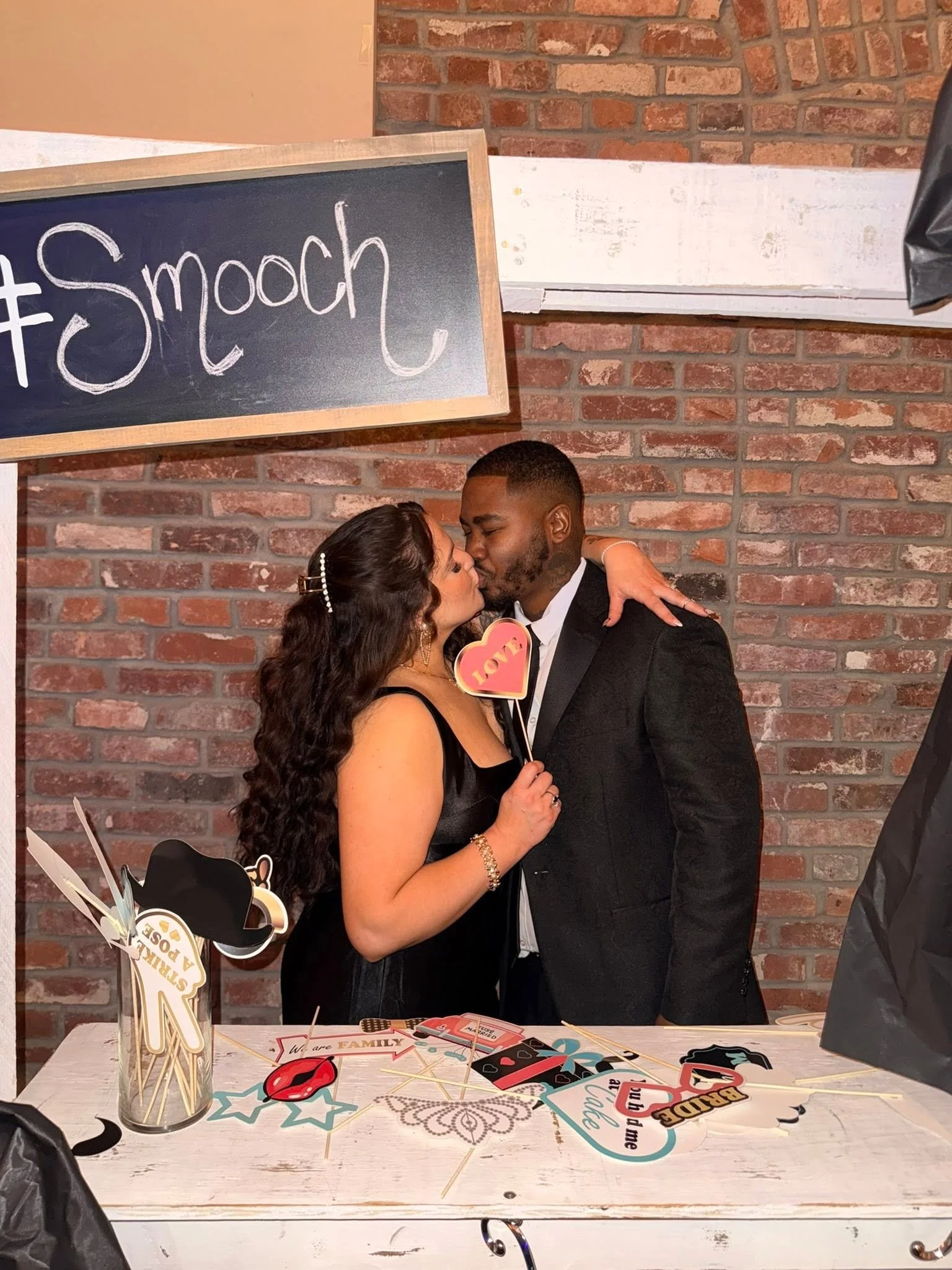 A couple sharing a kiss at a celebration table with photo props, against a brick wall and a chalkboard sign that says '#Smooch'.