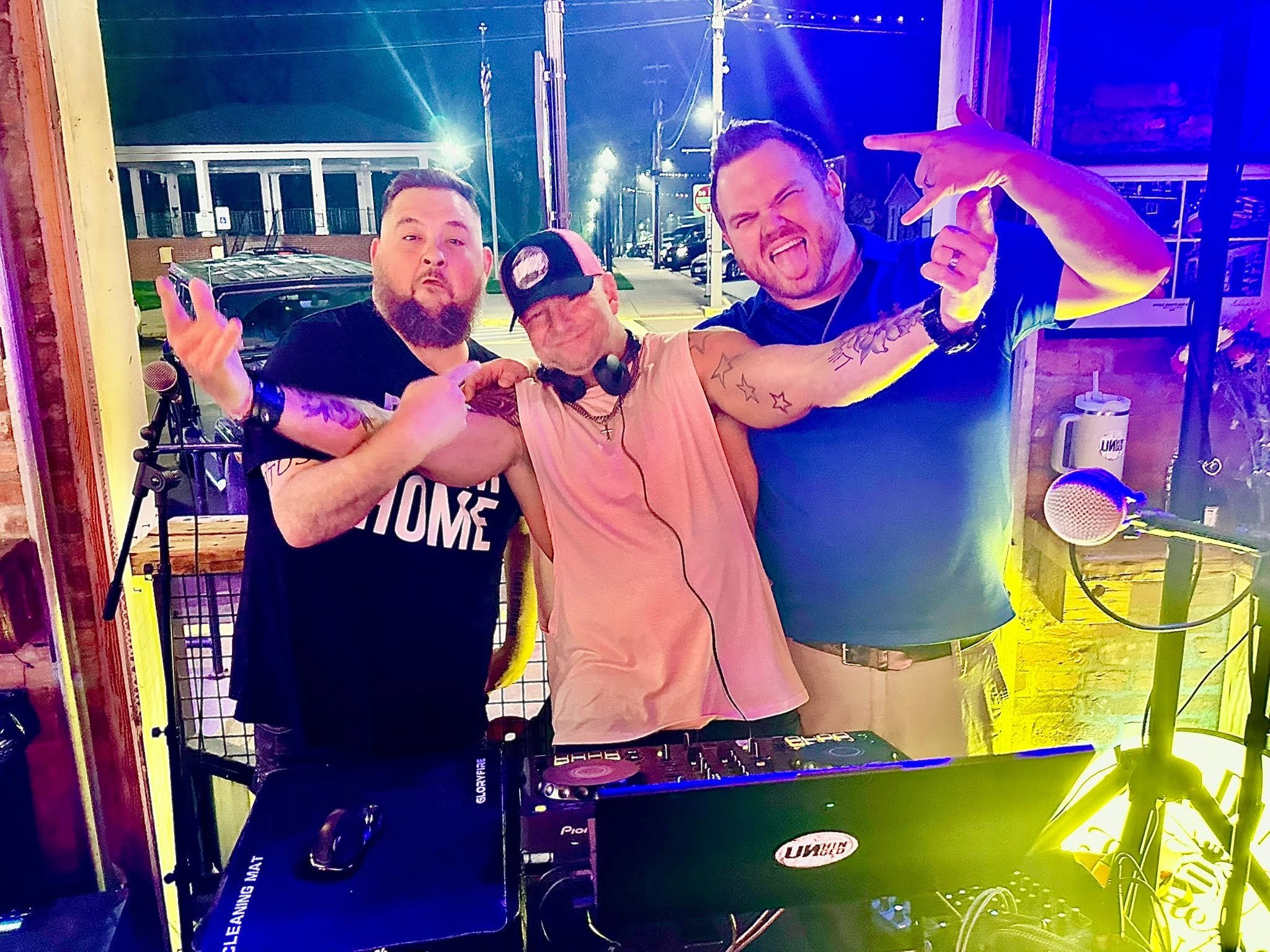 Three men are posing in front of DJ equipment outside at night. They are making hand gestures and sticking out their tongues, with colorful lights illuminating the scene.