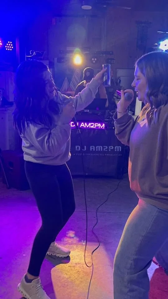 Two women singing into microphones at a karaoke night, with a DJ in the background and colorful stage lighting.