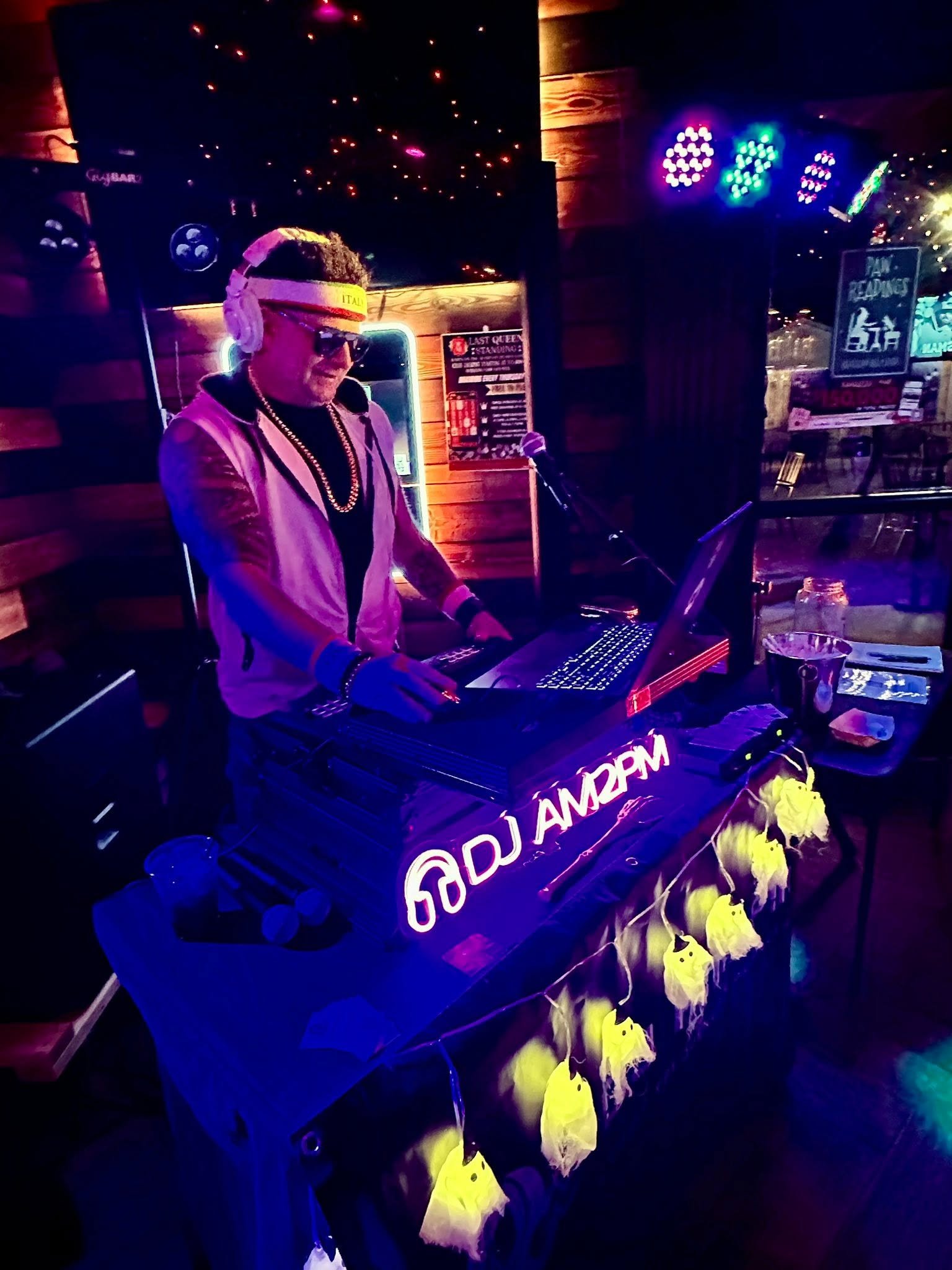A DJ wearing sunglasses, headphones, and a gold chain performs at a nightclub with colorful LED lights and a neon sign displaying social media handles.