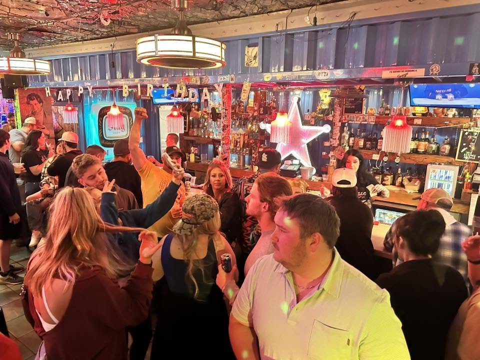 Crowded bar with people socializing and dancing, decorated with neon lights including a star, vintage posters, and a row of liquor bottles behind the bar counter.