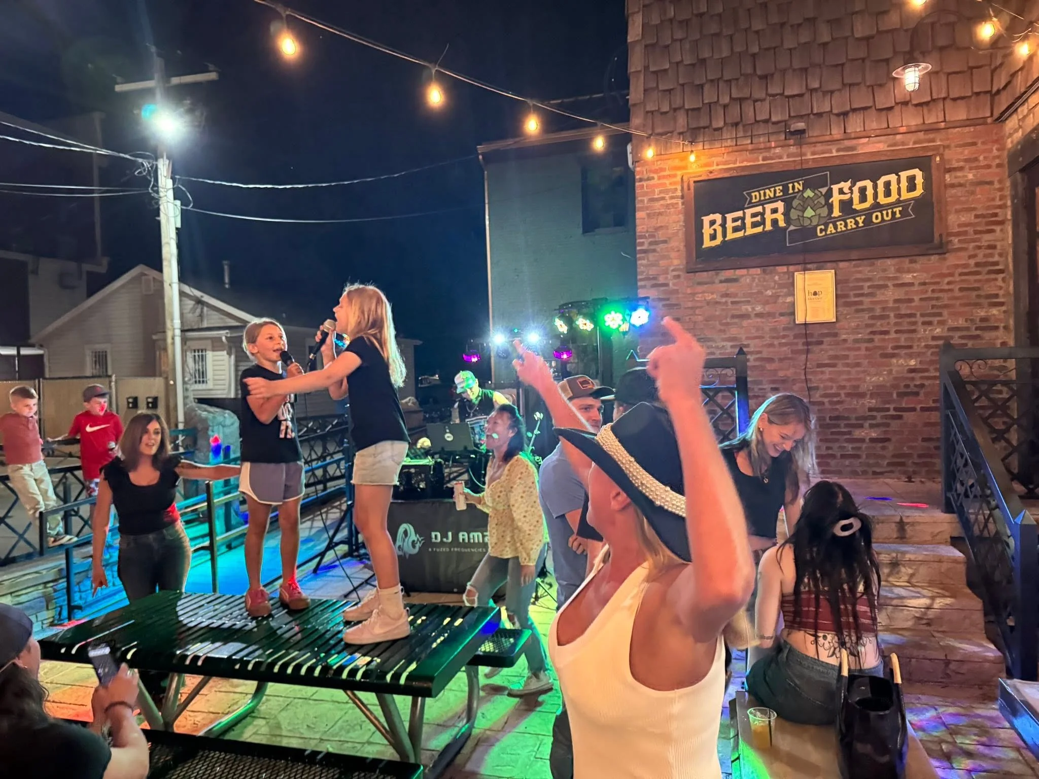 People dancing and enjoying live music at an outdoor nighttime event with string lights, a DJ playing music, and a sign for a beer and food patio.