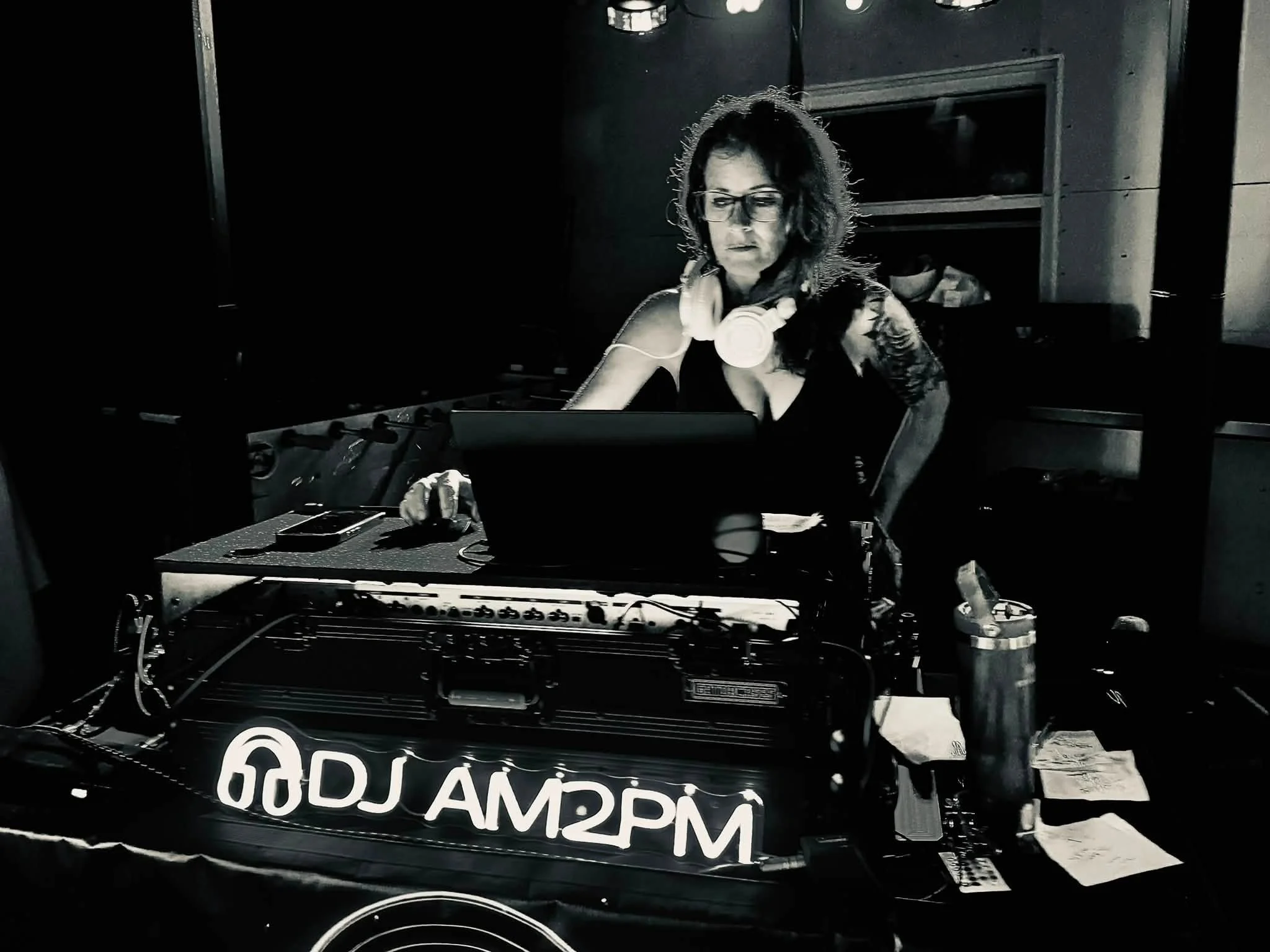 A woman wearing glasses working on electronic equipment with headphones around her neck, inside a dark room.