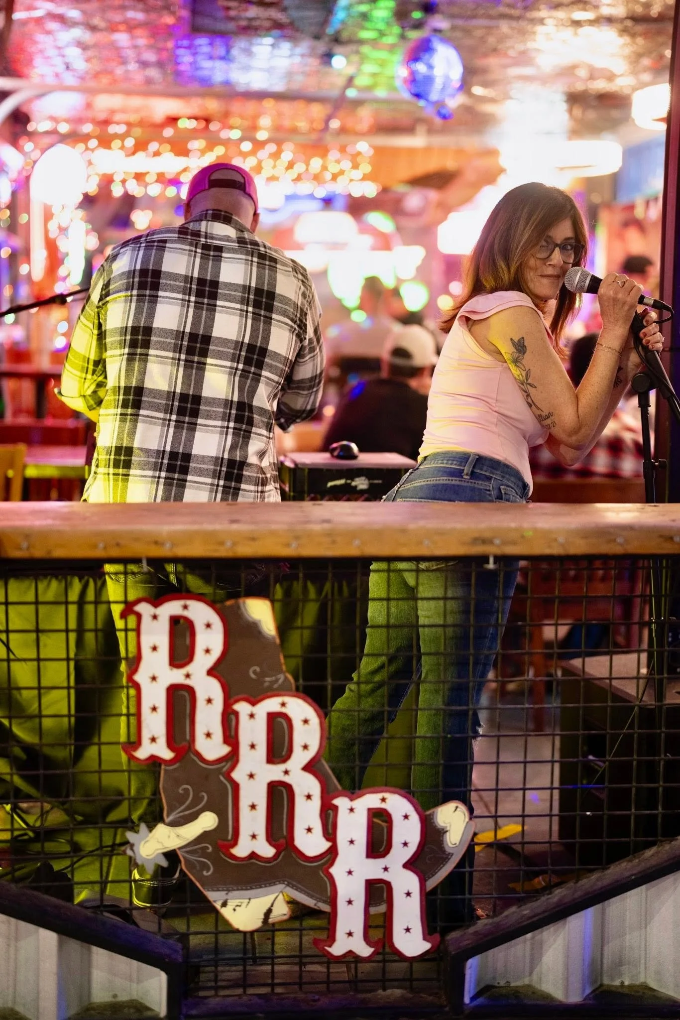 A woman with glasses holding a microphone, standing next to a man in a checkered shirt with his back turned. They are inside a festive venue with colorful lights and decorations, including a sign that says 'BIRD'.