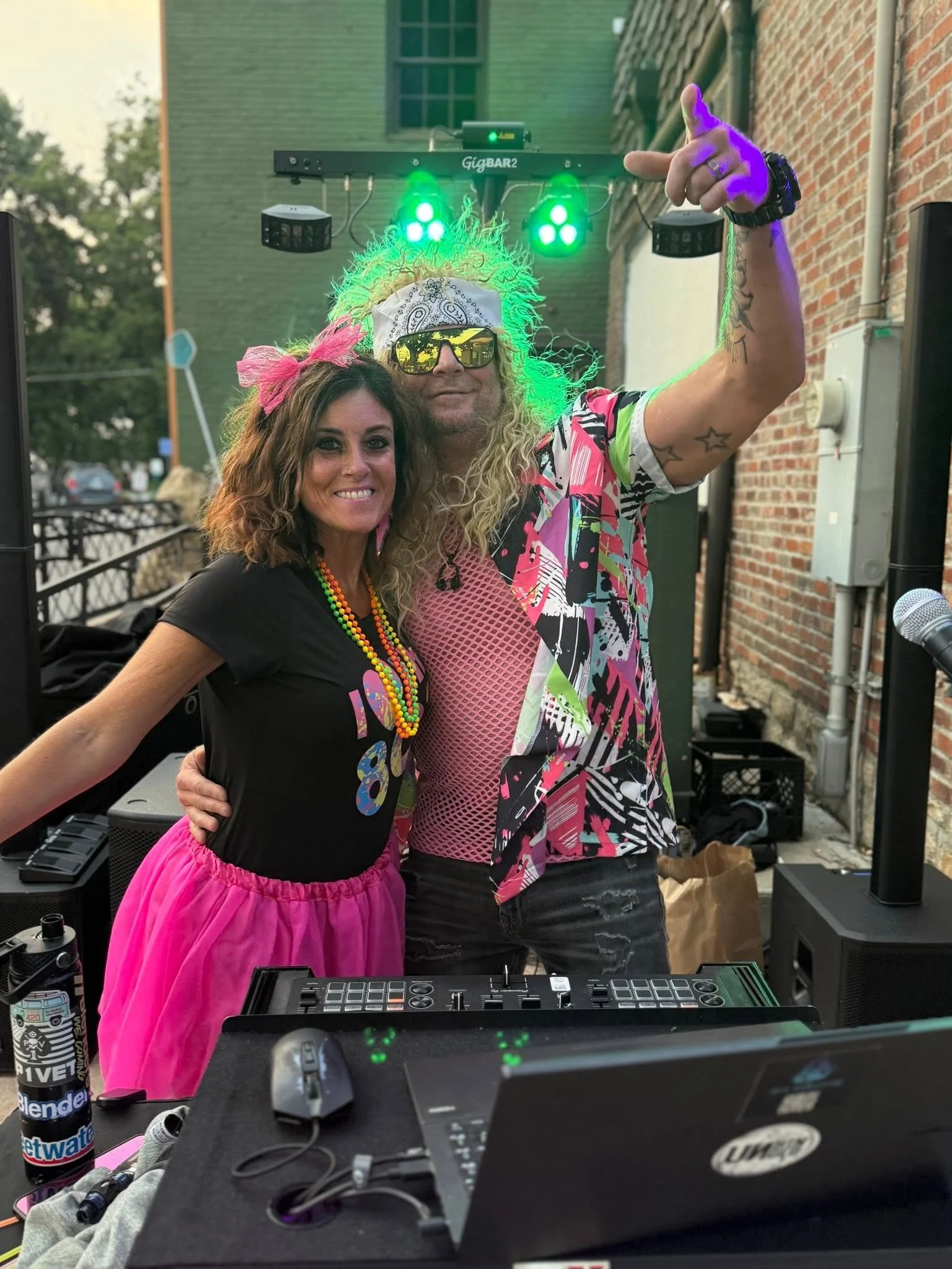 A woman with a curly hair styled with pink bows, wearing a black top, pink tutu, and colorful beaded necklaces, posing with a man with long curly blond hair and a beard, wearing sunglasses, a white bandana, a vibrant patterned shirt, and making a han