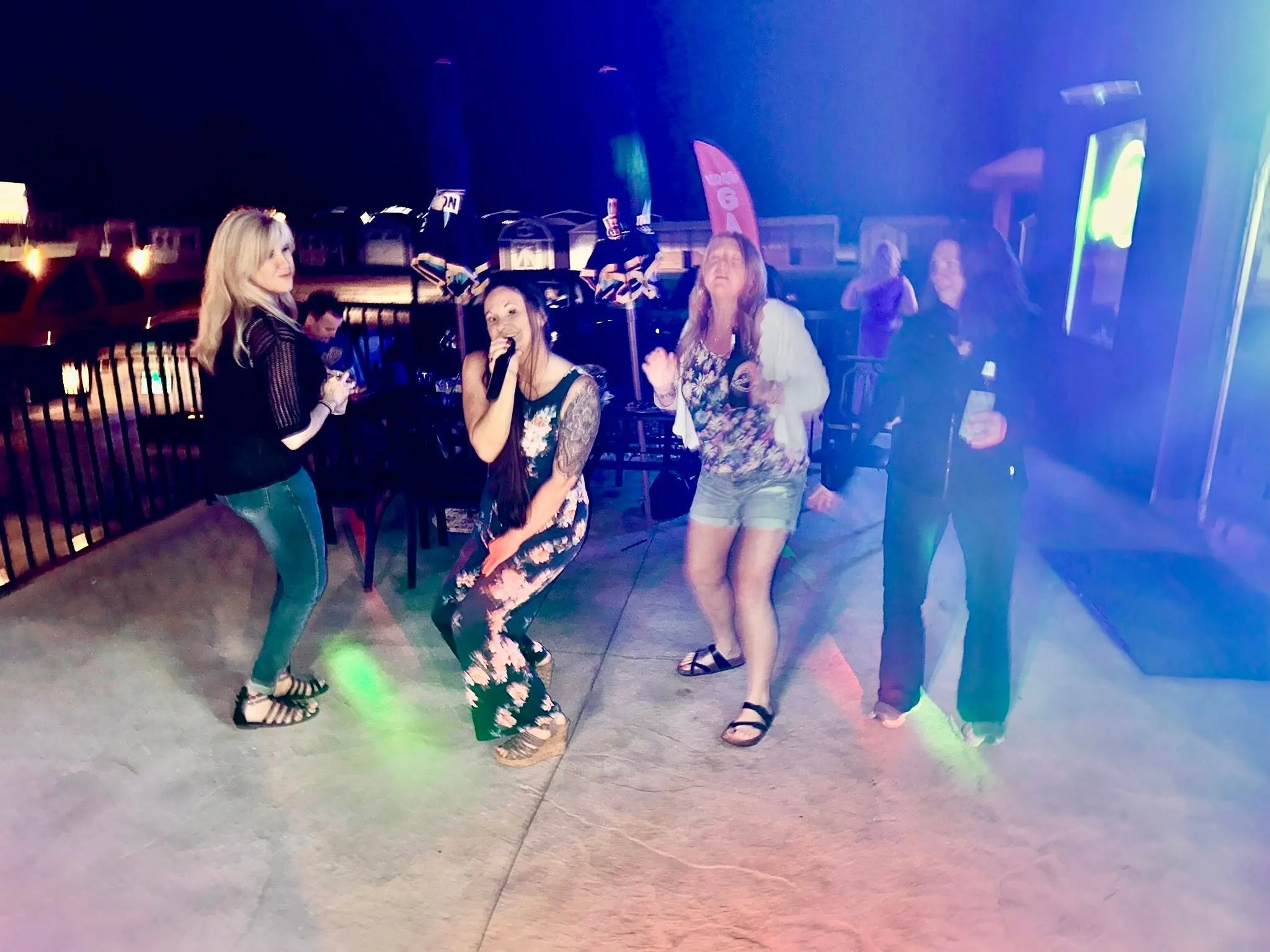 Four women are dancing and enjoying themselves outdoors at night, with colorful party lights, near a fence and city buildings.