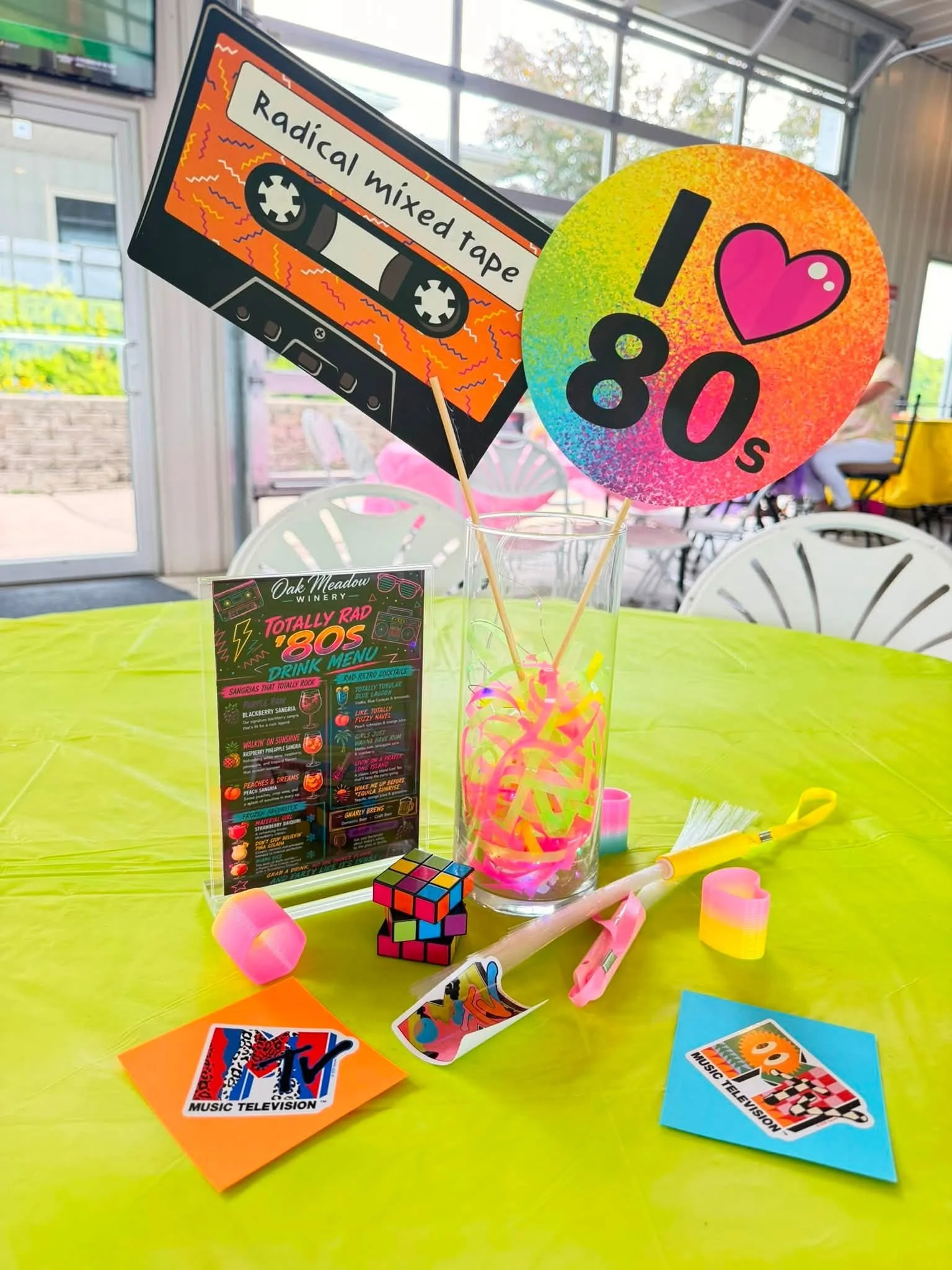 Colorful table decorated with neon pink, orange, and blue decorations, posters, and paper items celebrating the 1980s music and TV themes. Includes a Rubik's cube, a glass vase with neon ribbons, a Burger King sticker, and 80s themed signs that say "