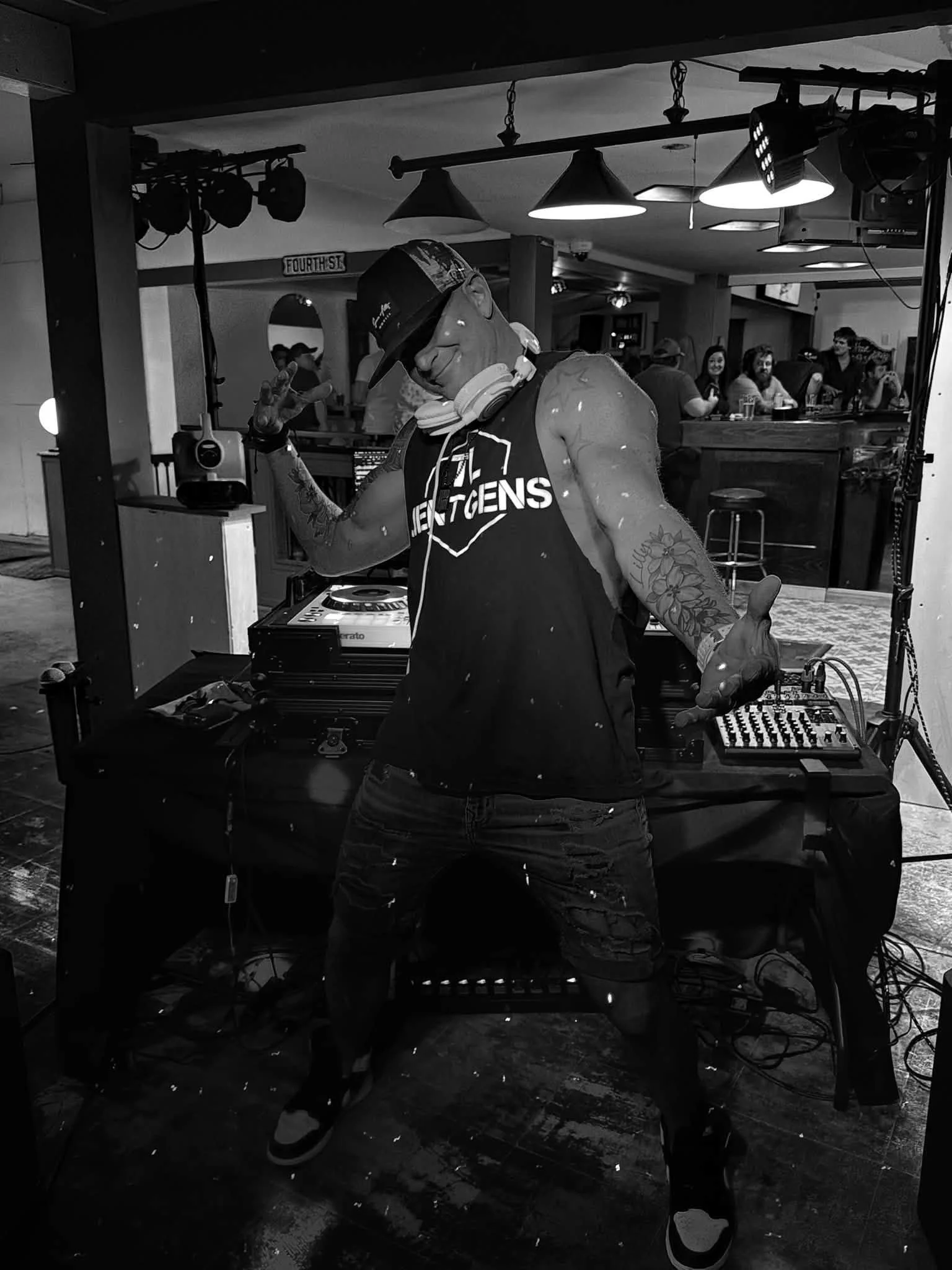 A DJ wearing a black tank top with the word 'LENGTHENS,' headphones, and a cap is performing at a bar or club, with people sitting and standing in the background.