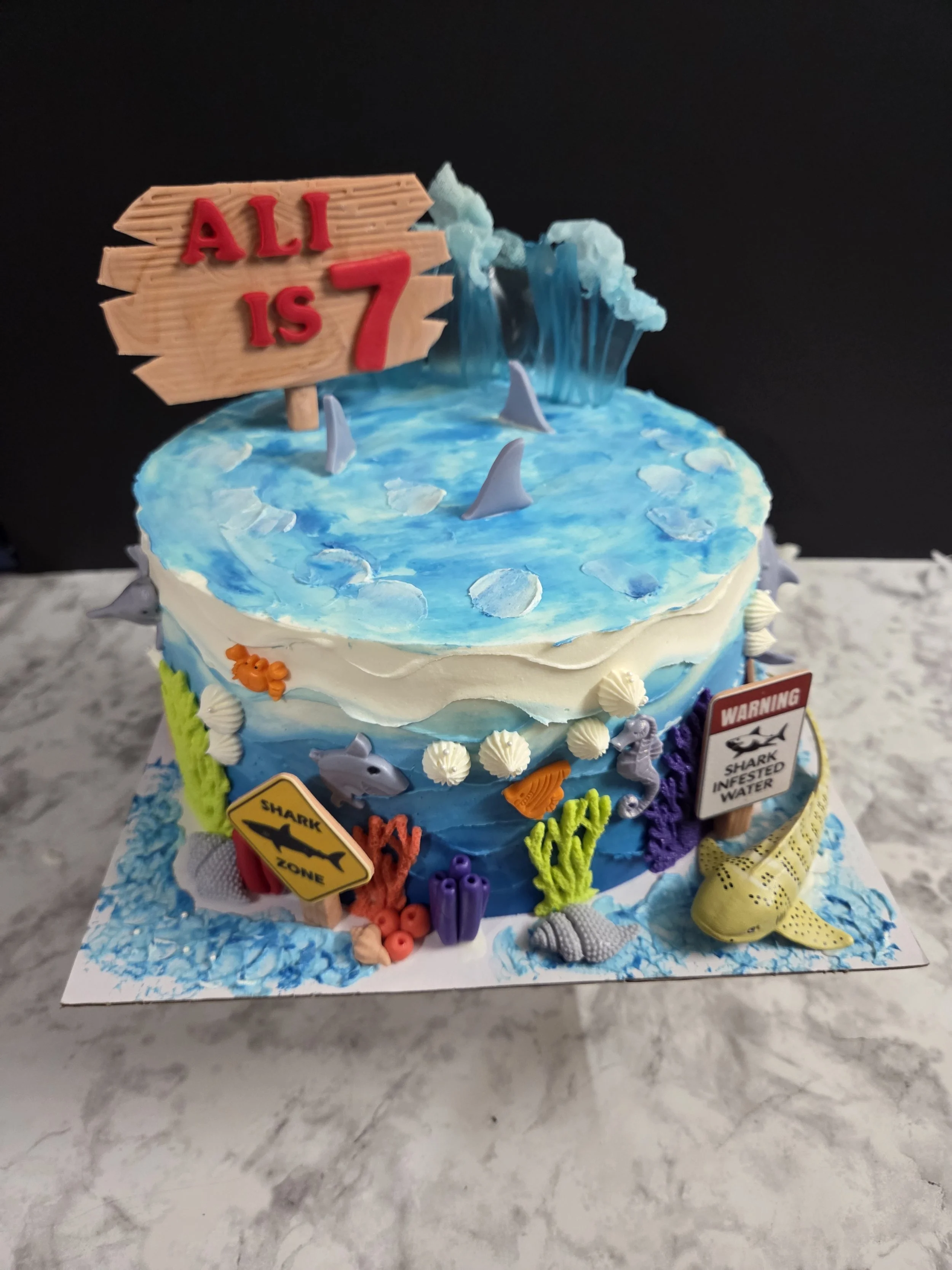 Custom ocean-themed 7th birthday cake featuring 100% edible decorations, blue ombre ocean wave icing, shark fins, fondant coral reef accents, a fondant wooden sign, and sugar sculpted waves, handcrafted at Sloan's Sweets and Sammies in Myrtle Beach.