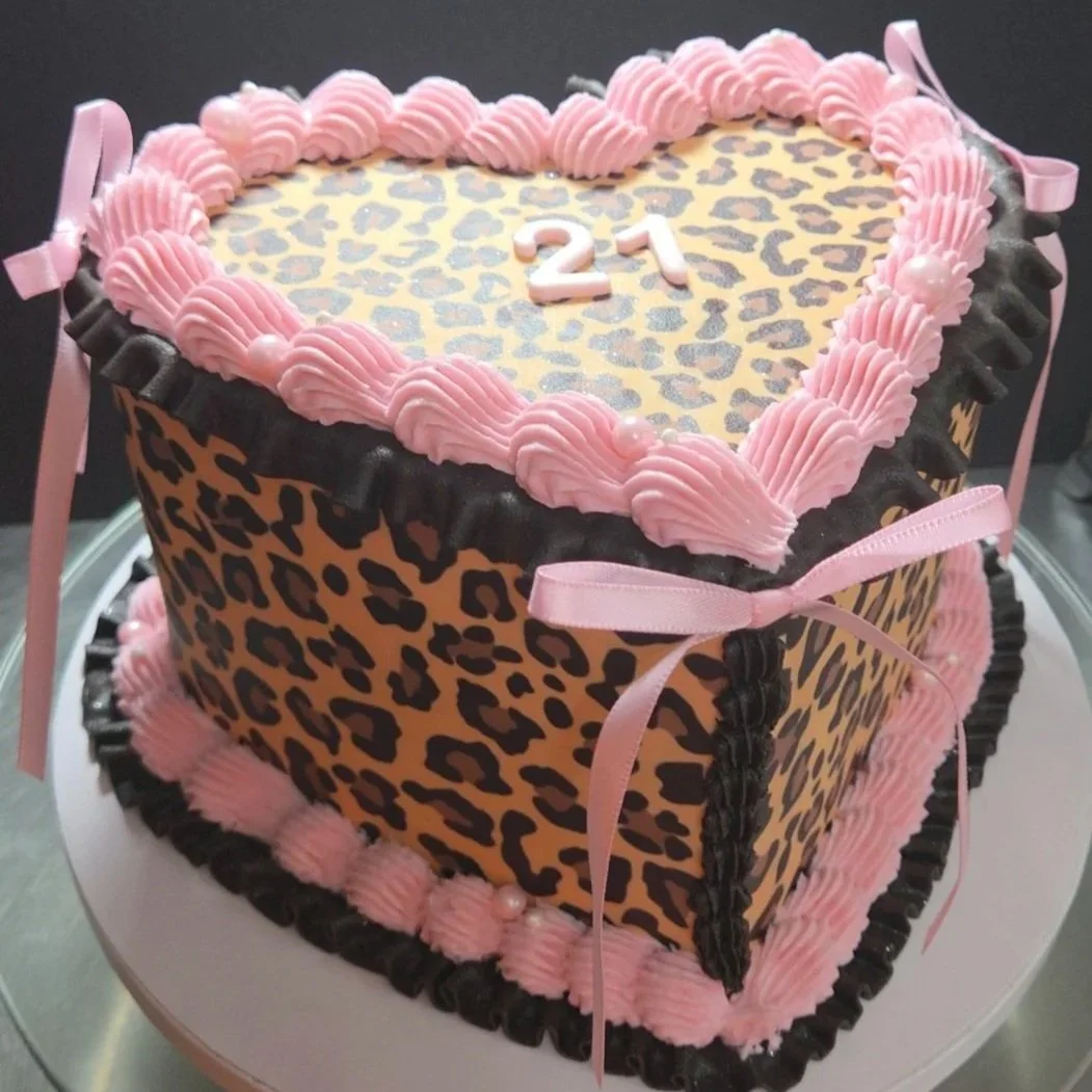 A heart-shaped cake decorated with leopard print, pink and black icing, pink ribbons, and small pink pearls, with the number 21 on top.
