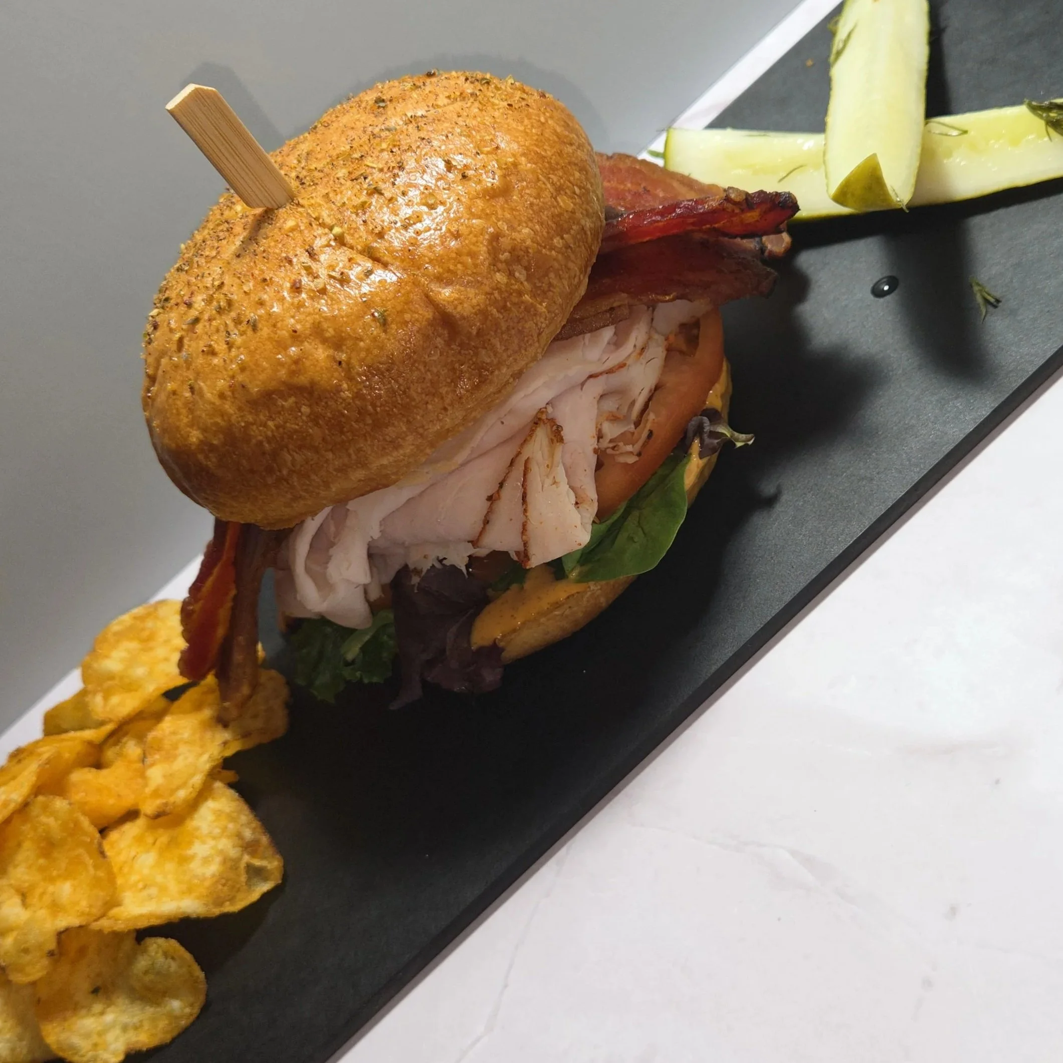 A Turkey Bacon Chipotle Ranch sandwich stacked with thinly sliced turkey, crispy bacon, spring mix, and tomato on a toasted kaiser roll. Served on a black platter, accompanied by sliced pickle and barbeque chips.