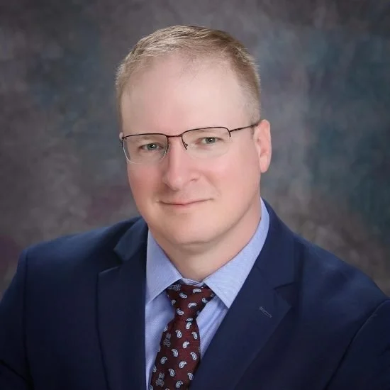 A man with glasses, wearing a navy blazer, light blue shirt, and patterned tie, posing against a textured background.