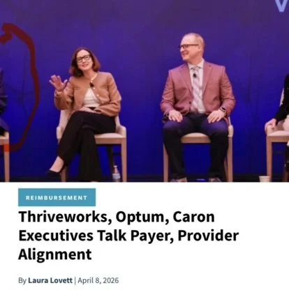 Three people sitting on chairs on a stage participating in a panel discussion with a blue background. The woman on the left is wearing glasses and a light brown blazer, smiling and gesturing with her hands. The man in the middle is wearing glasses, a pink blazer, and a white shirt, smiling with his hands on his lap.