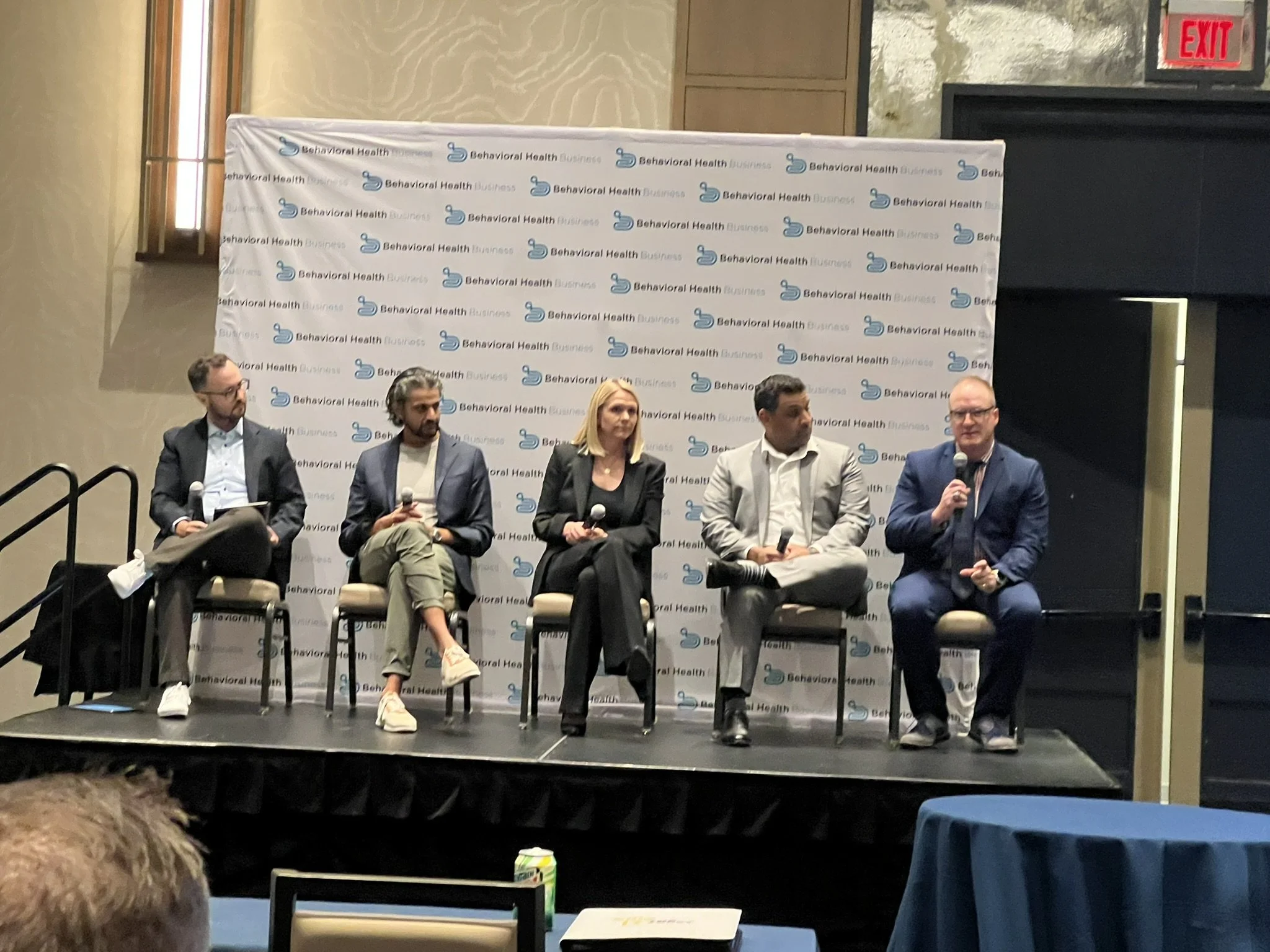 A panel discussion at an event with five individuals seated on stage, microphone in hand, in front of a branded backdrop with the logo and text 'Behavioral Health Business'.