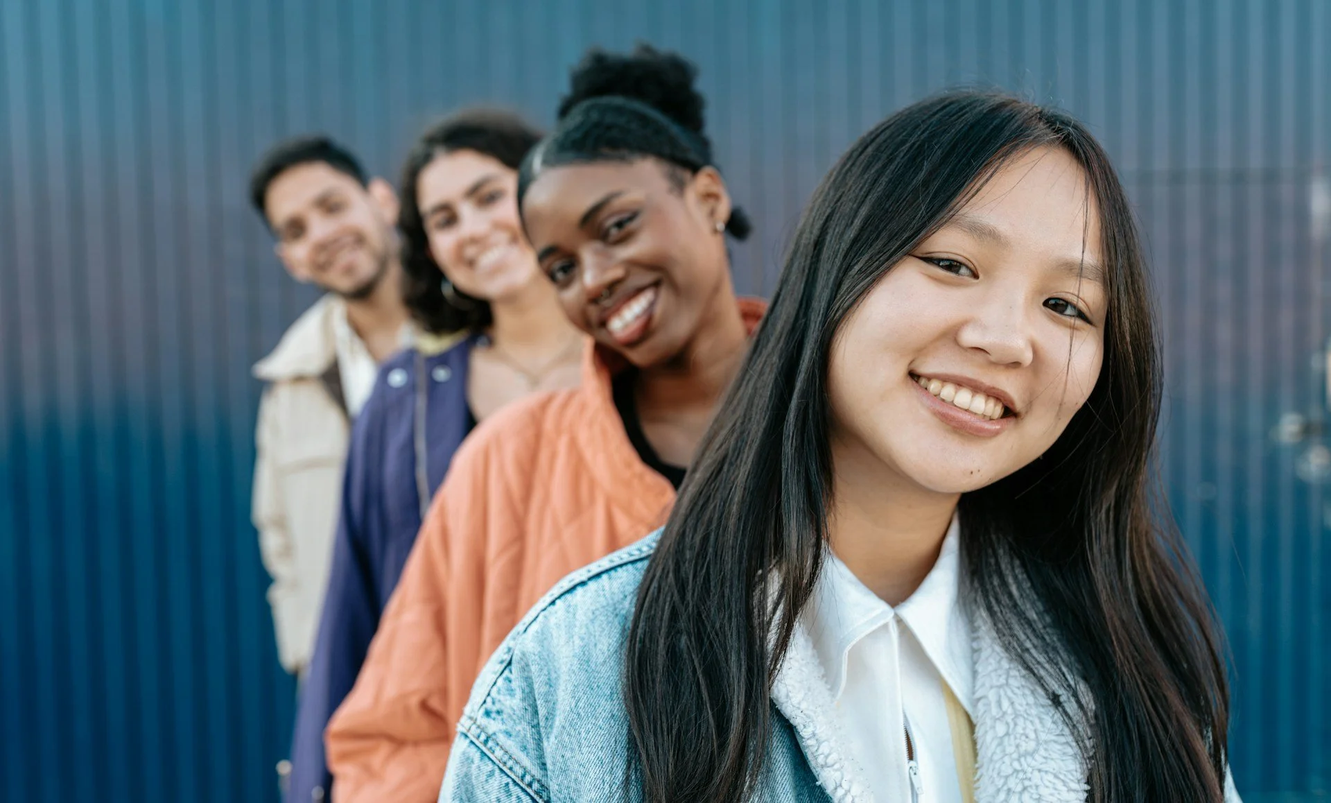 Group of four diverse young people smiling in a line against a blue background.
