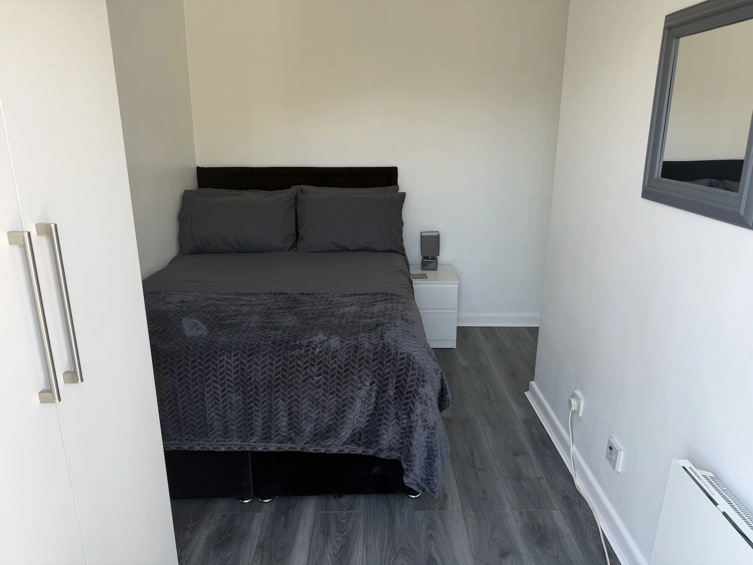 A bedroom with a bed, gray bedding, a nightstand with a small speaker and a phone, a mirror on the wall, and a closet.