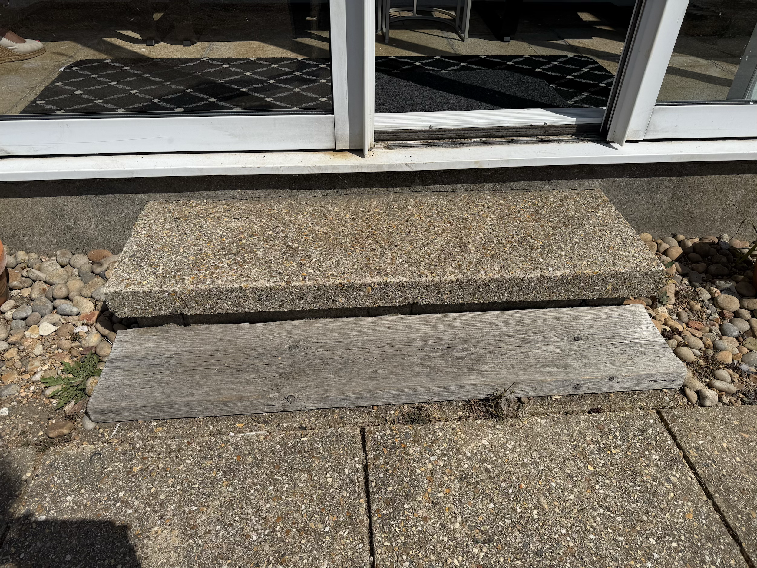 Glass door with a black patterned rug inside, concrete step with wood and brick pavements, and pebbles in the foreground.