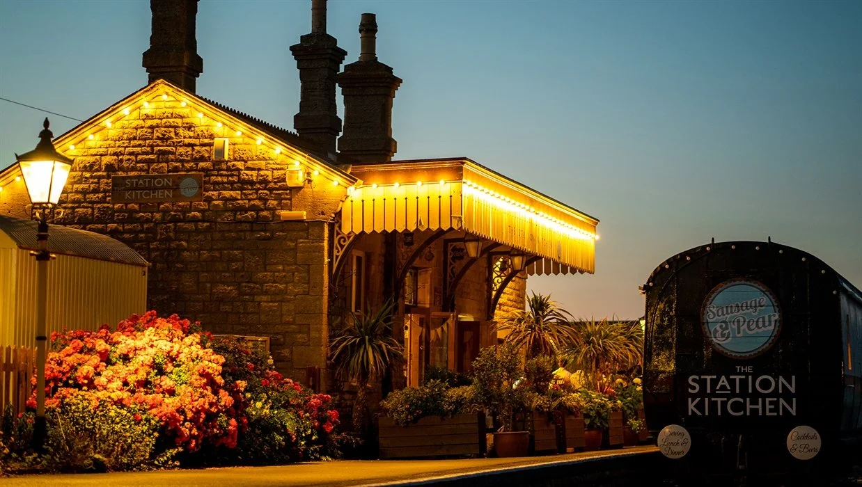 A rustic building at dusk with illuminated string lights, surrounded by colorful flowers and potted plants, and a vintage street lamp in the foreground. A black wagon with sign fronts the scene.