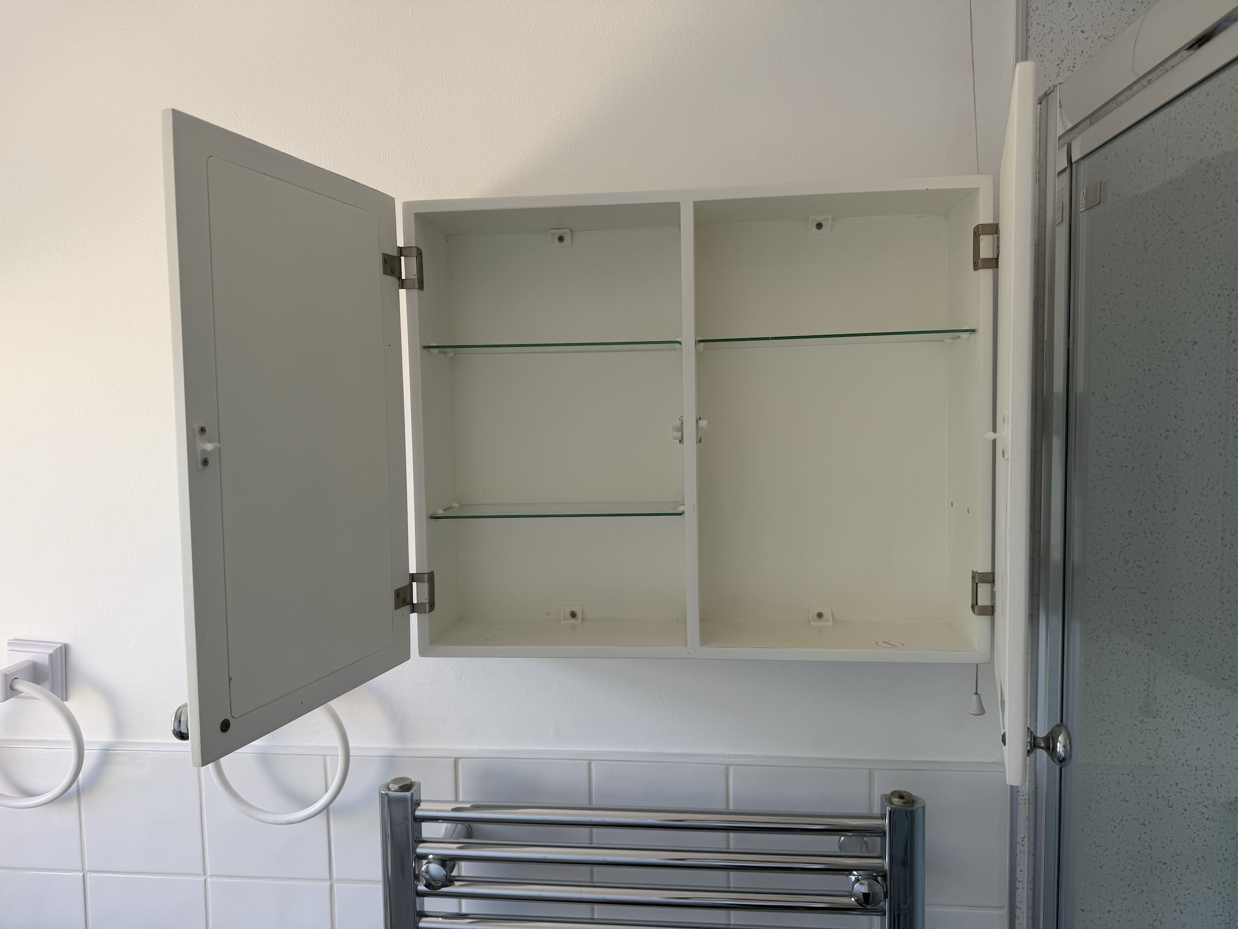 Empty open white bathroom medicine cabinet with glass shelves, mounted on a white wall, with a small hook at the bottom and a mirror on the outside of the door.