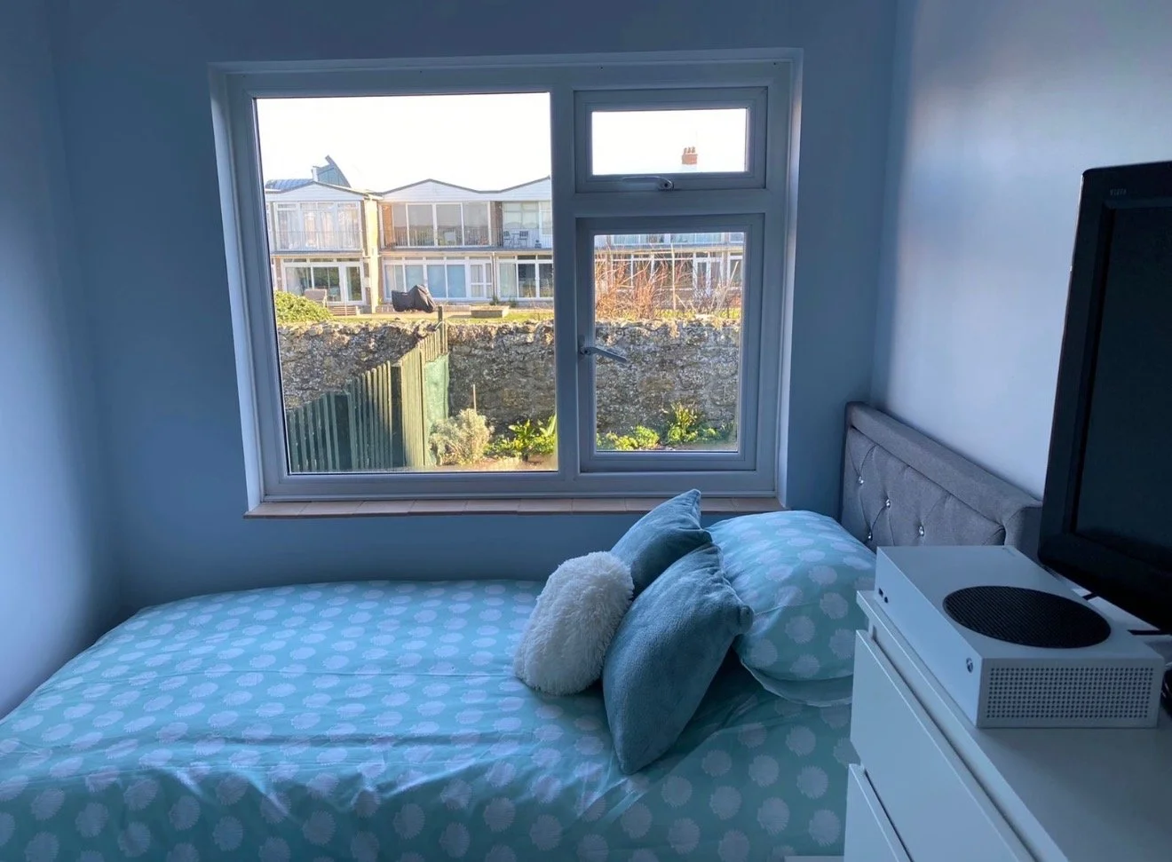 A bedroom with a bed covered in blue polka dot bedding and pillows, positioned near a large window showing neighboring houses and a garden outside.