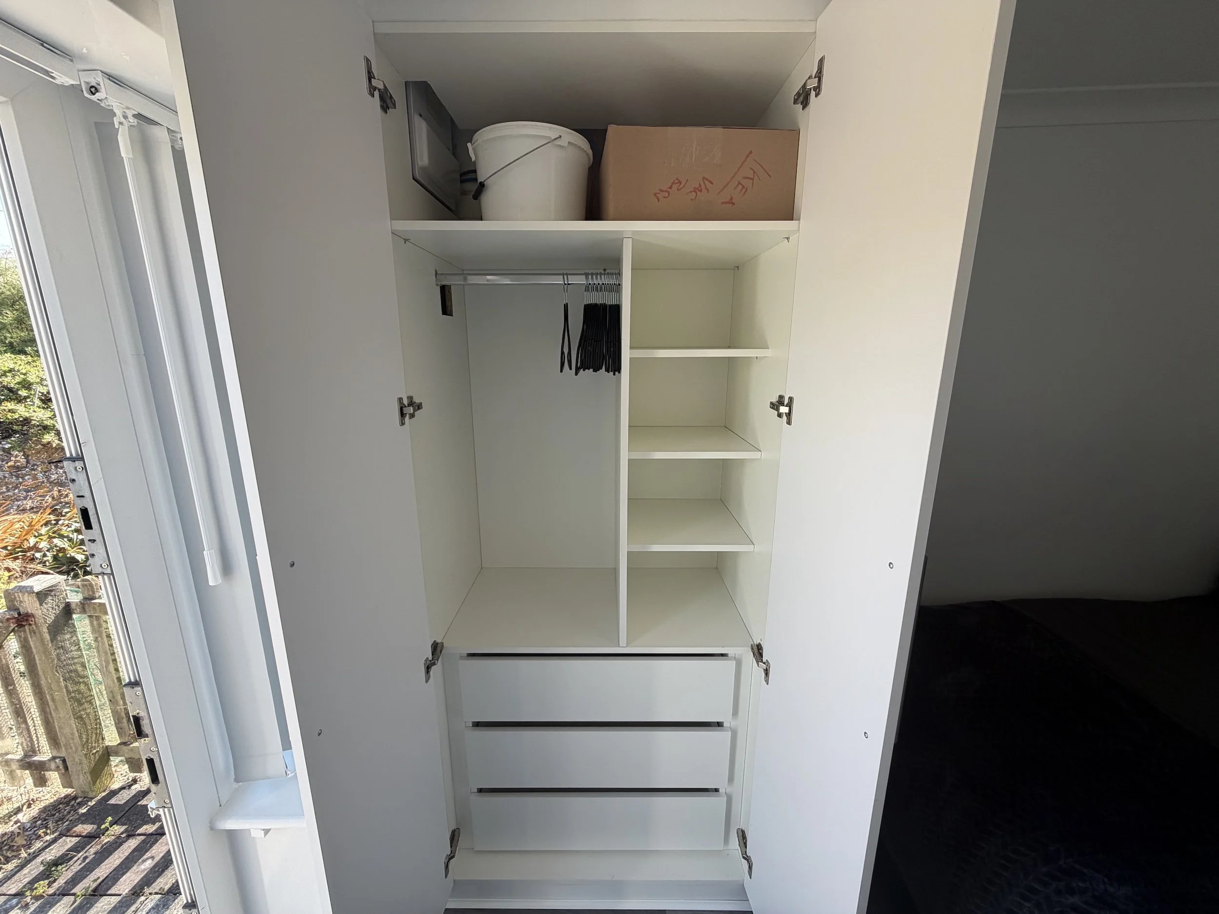 Empty white kitchen cabinet with open doors, showing shelves with some items including a white bucket, a cardboard box with writing, and black hangers.