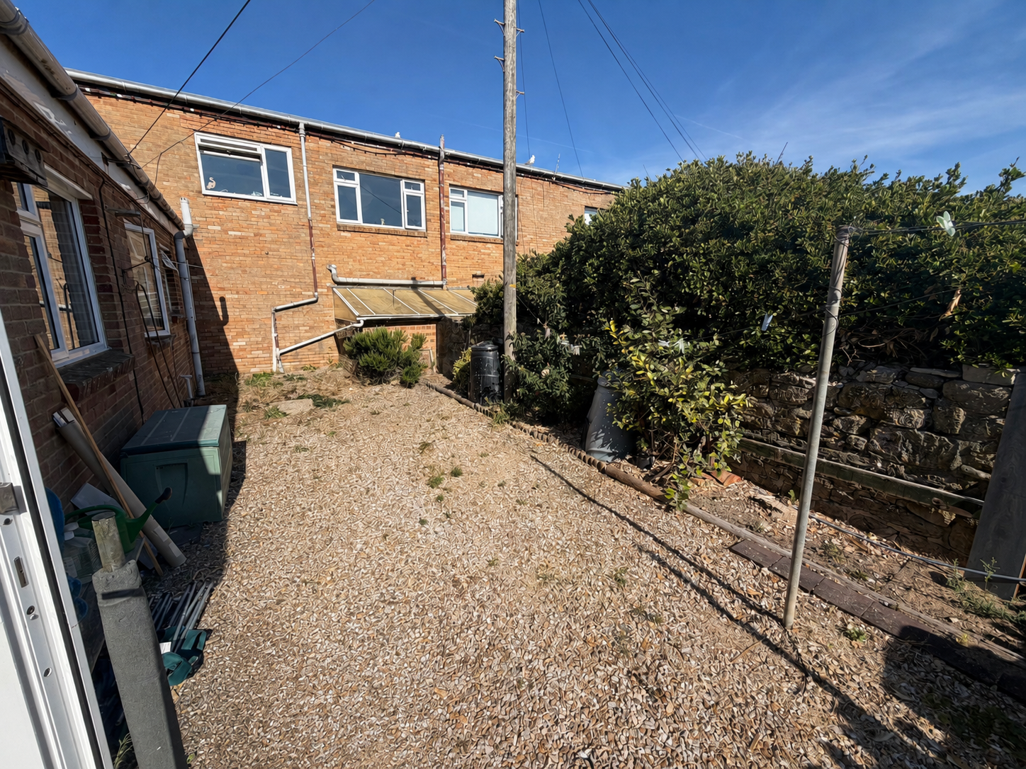 Small backyard with gravel ground, a green garden storage box, various gardening tools, and a dried-up shrub. A telephone pole with wires is present, and there is a large green bush on the right. Brick houses with windows are in the background under 