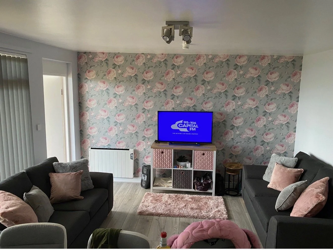 Living room with floral wallpaper accent wall, two dark sofas with pink and gray pillows, a TV on a pink and white cubed media stand, a small pink rug, and a heater on the wall.