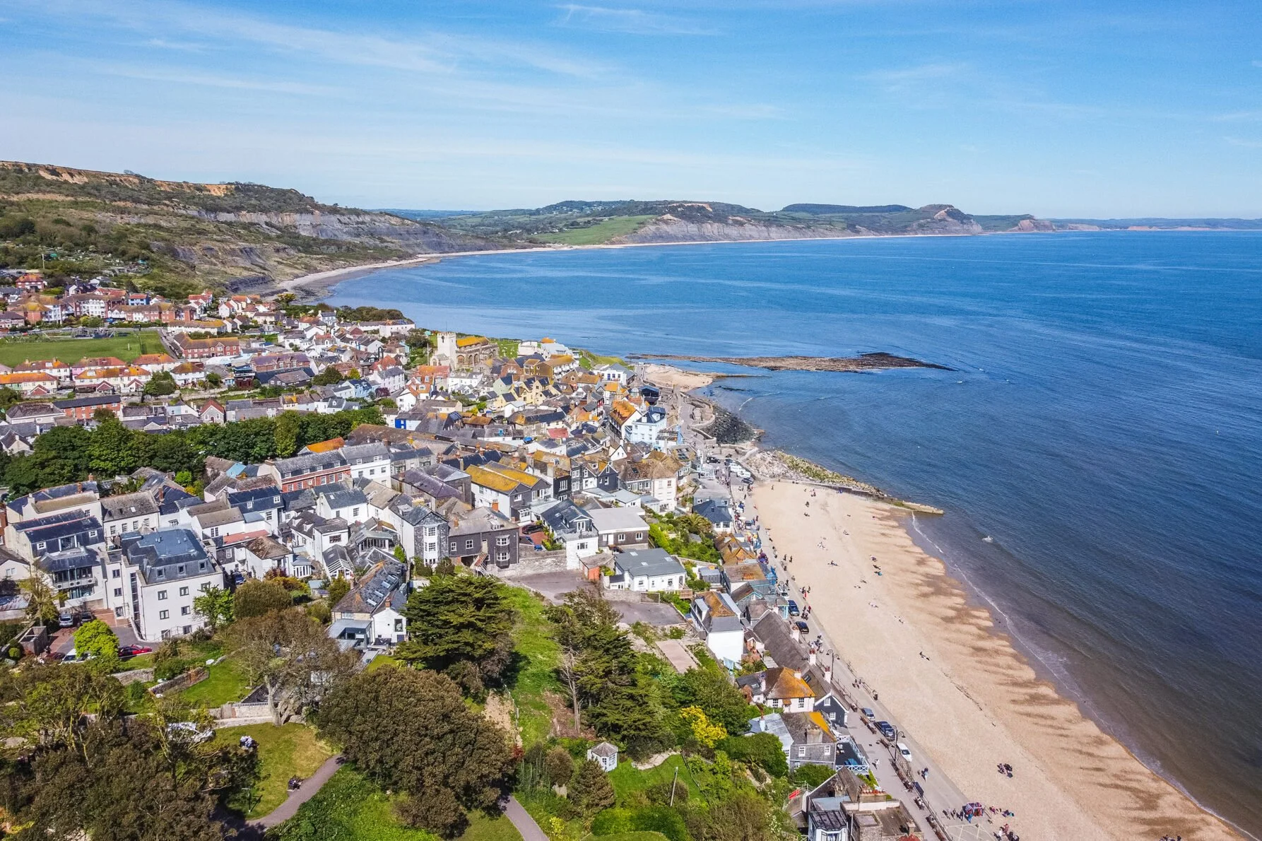 A coastal town with colorful houses along the shoreline, a sandy beach, and cliffs in the background under a partly cloudy sky.
