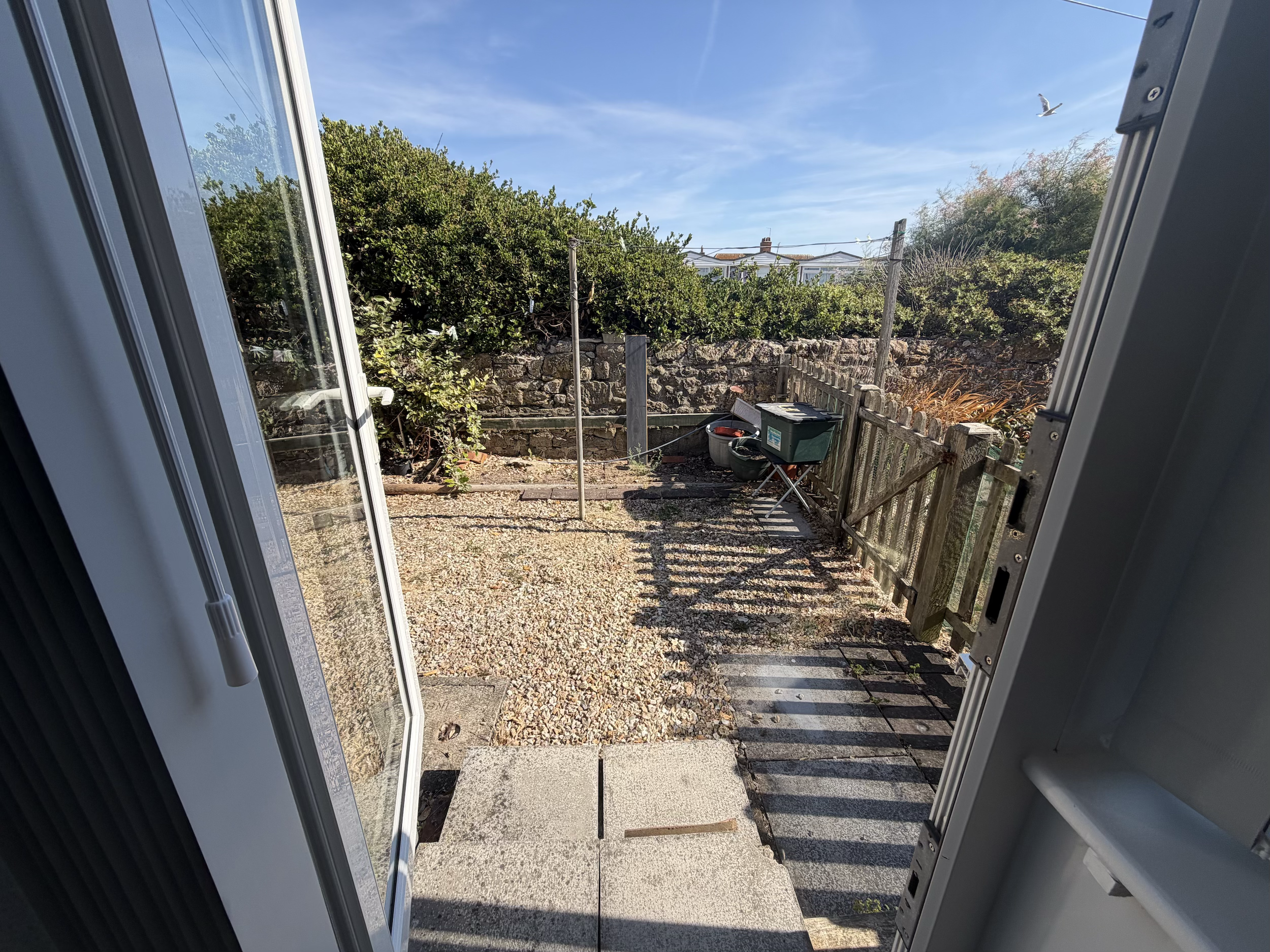 View through an open door showing a backyard with a gravel path, a small concrete patio, gardening supplies, a green fence, and trees in a sunny scene.