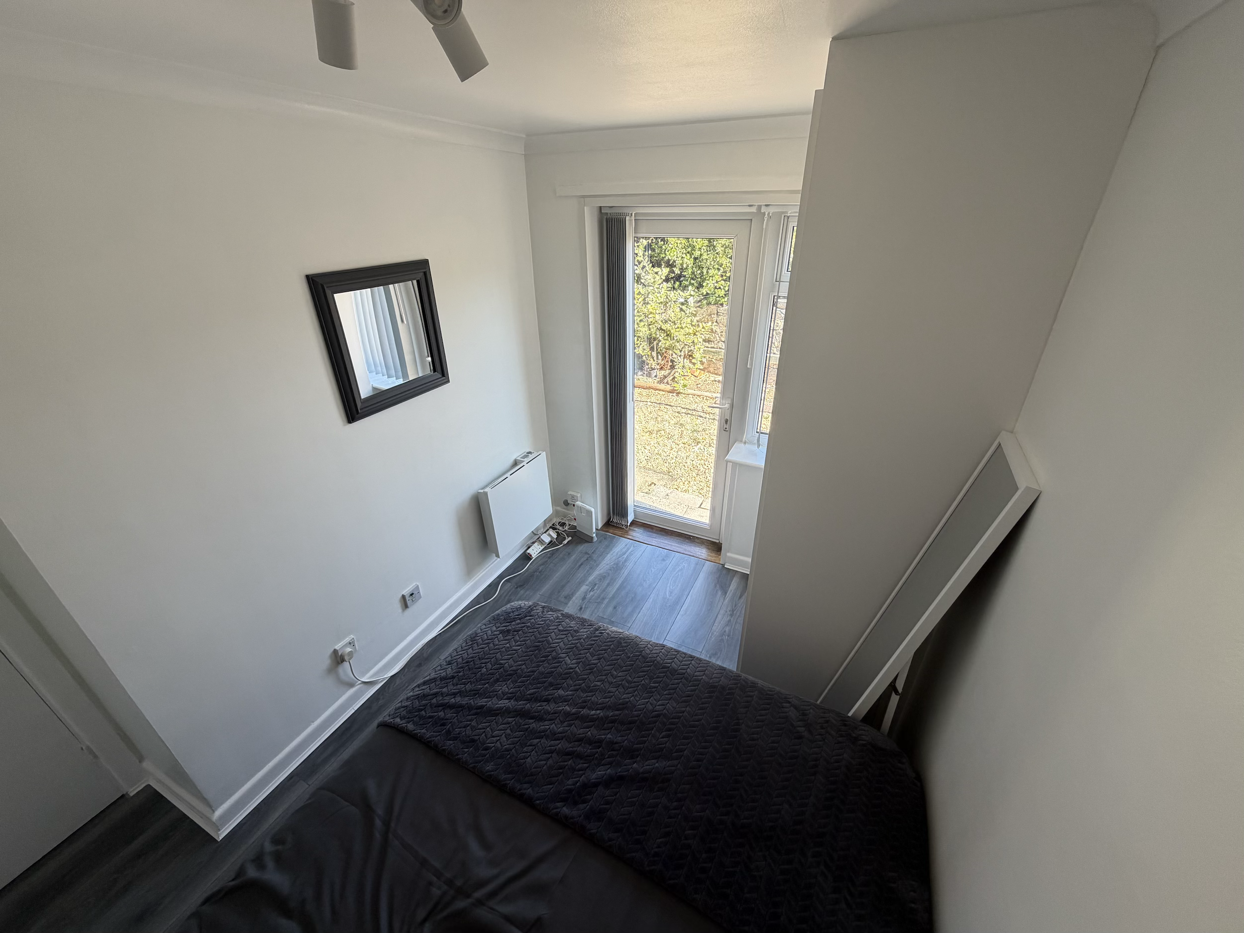 A small room with white walls and a dark wood floor, featuring a window with vertical blinds open to a view of trees outside. A mirror is mounted on the wall, and a black quilted blanket covers part of a piece of furniture in the foreground. There ar