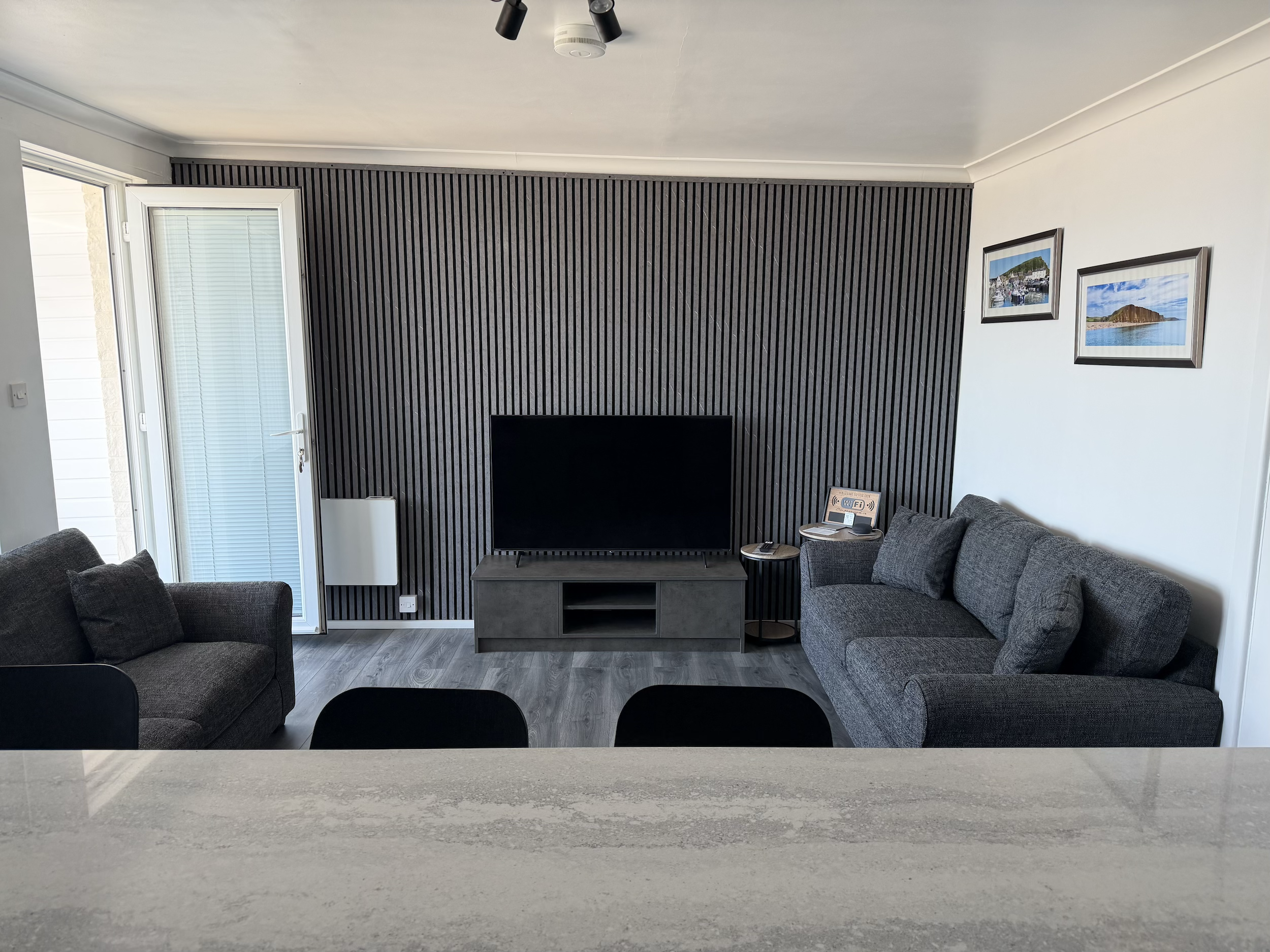 Living room with black wood-slatted wall, mounted flat-screen TV, gray sofas, framed seaside pictures, partially open glass door, wooden floor, ceiling with black and white light fixtures, Wi-Fi router on side table.