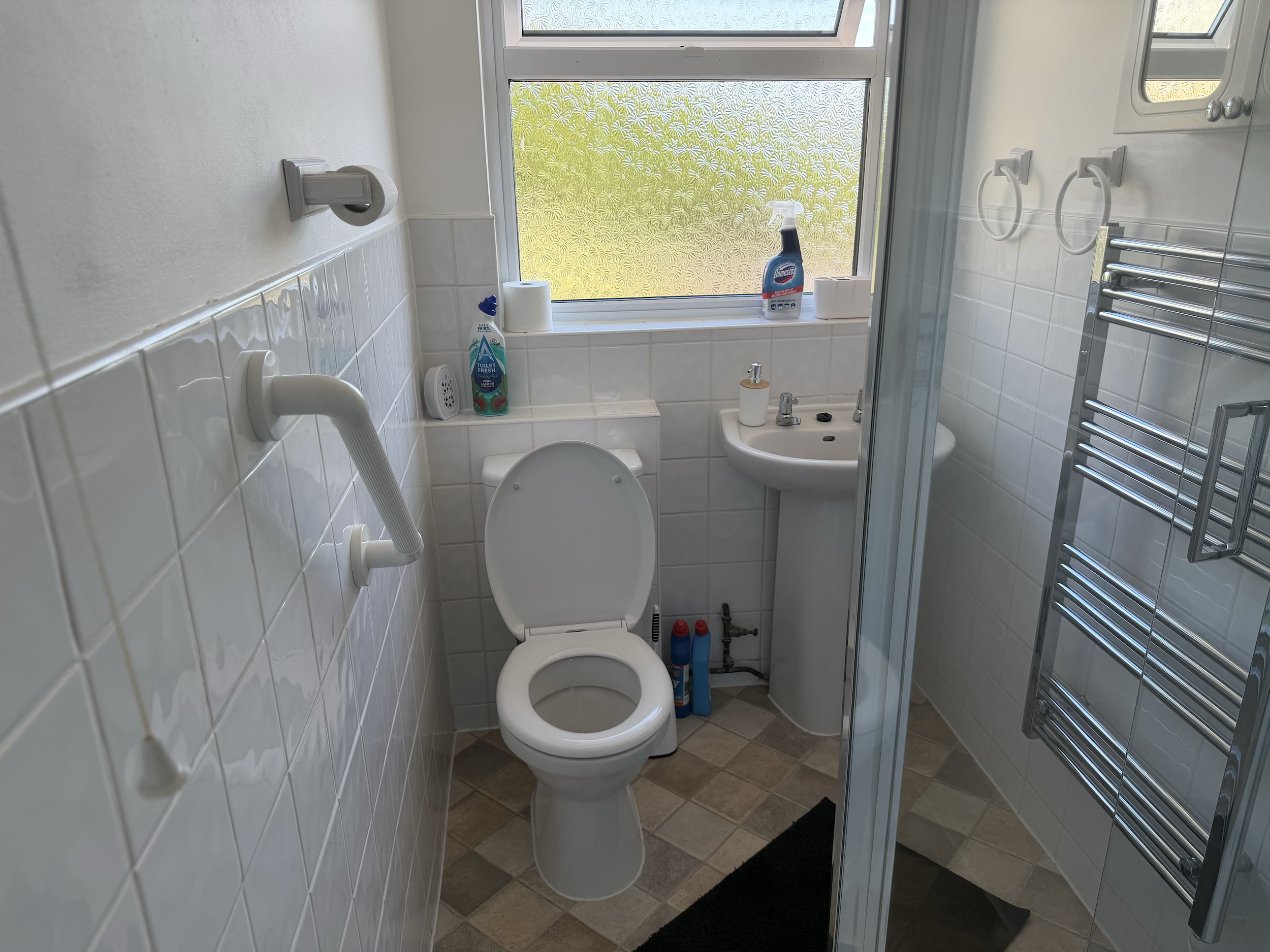 Small bathroom with white tiled walls, a toilet, a small sink, a frosted window, and various cleaning supplies on the windowsill. There are towel rings on the wall and a heated towel rail on the outside of the shower enclosure.