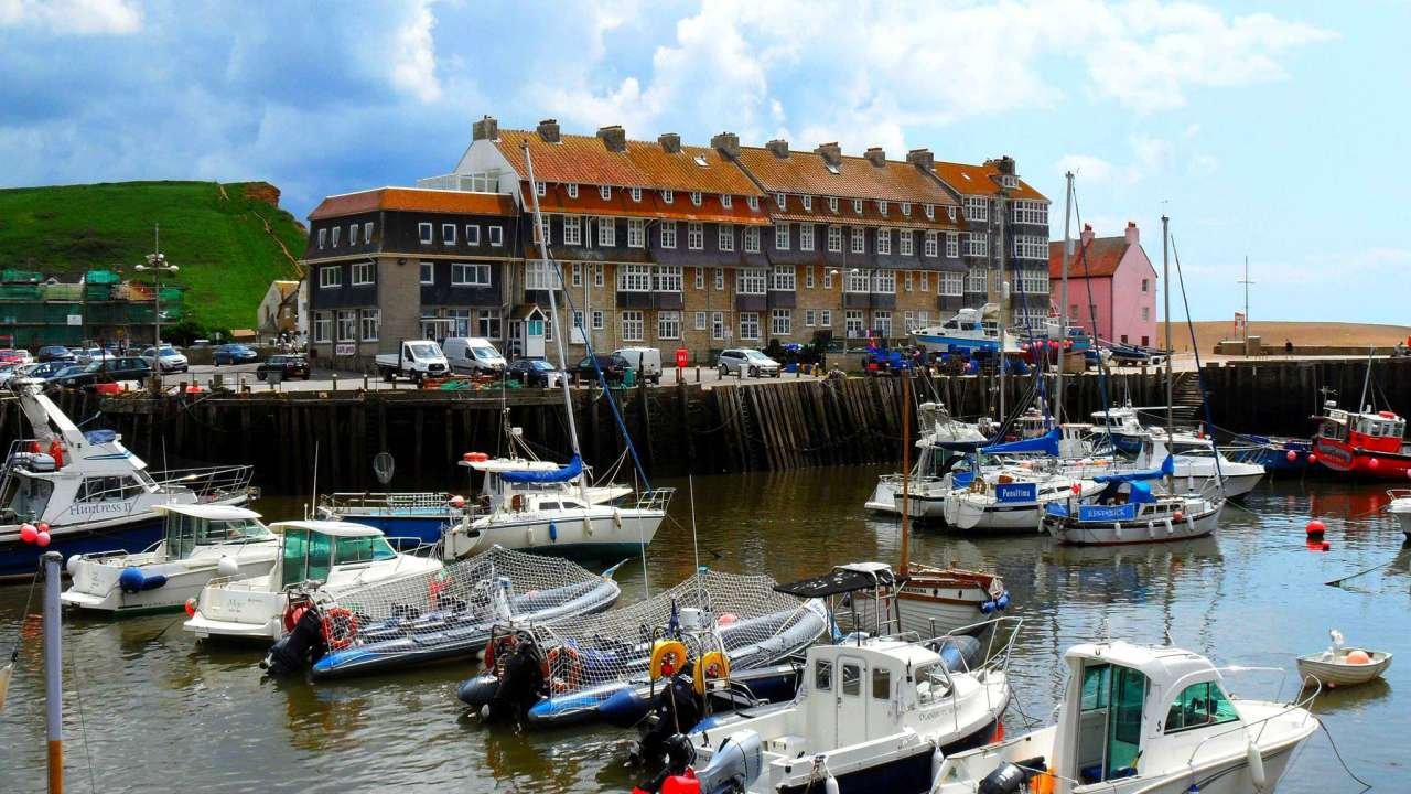Boats docked in a marina with colorful buildings and a hill in the background.