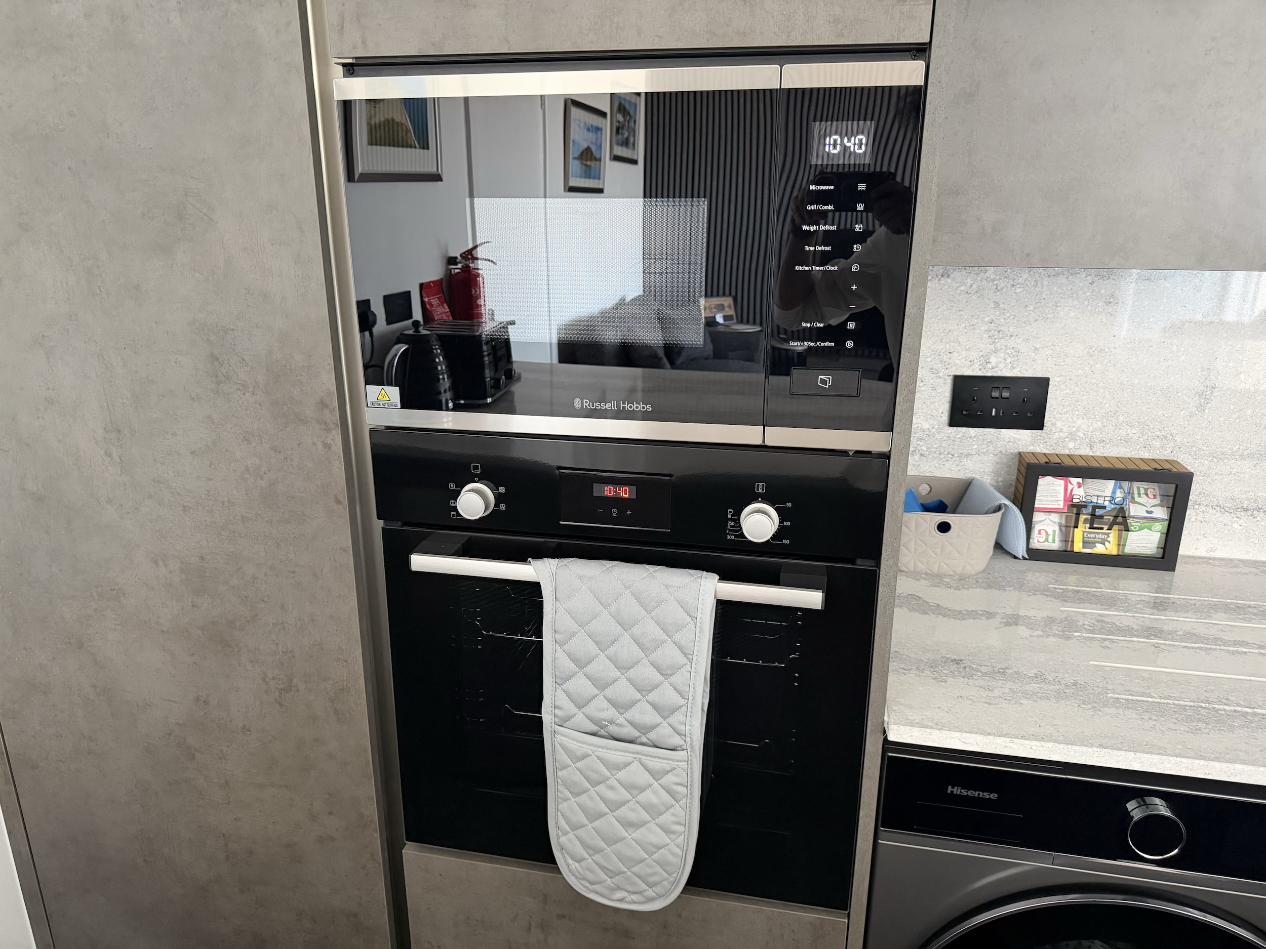 A kitchen microwave oven with digital controls, a glass door, and a white quilted oven mitt hanging on its handle, placed on a kitchen island or counter.