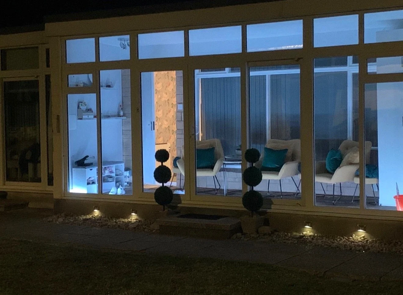 a sunroom or enclosed porch with large windows, white chairs with teal pillows, indoor lighting, and decorative black spheres on the floor