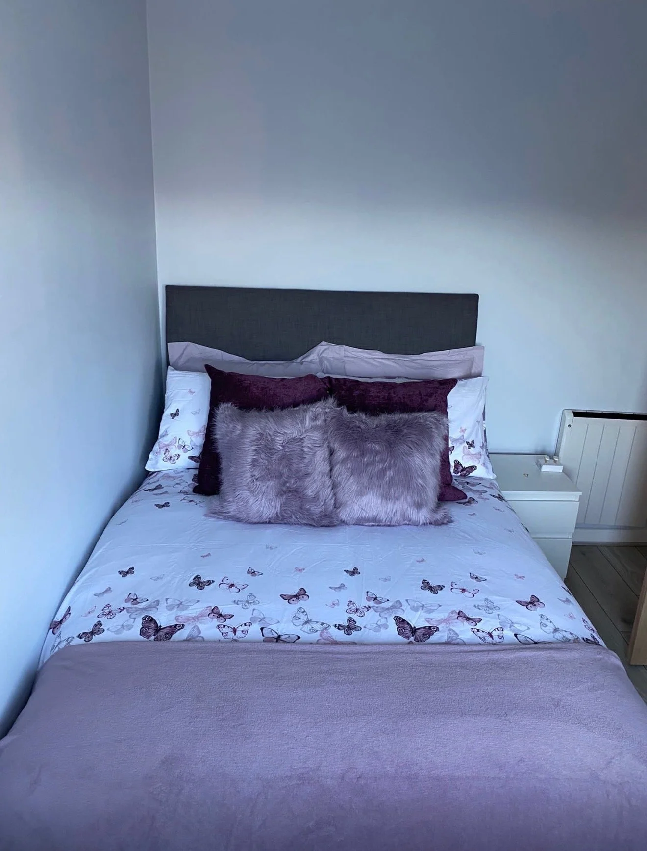 A neatly made bed with a butterfly-printed bedsheet, decorated with multiple pillows including a furry gray pillow, situated against a wall with a headboard and a small bedside table with a drawer and a heater nearby.