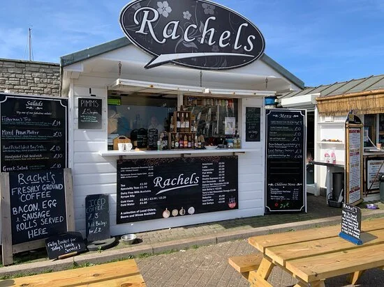 Small outdoor cafe stand named Rachel's selling coffee, snacks, and ice cream with menu boards, seating area, and a clear sky in the background.