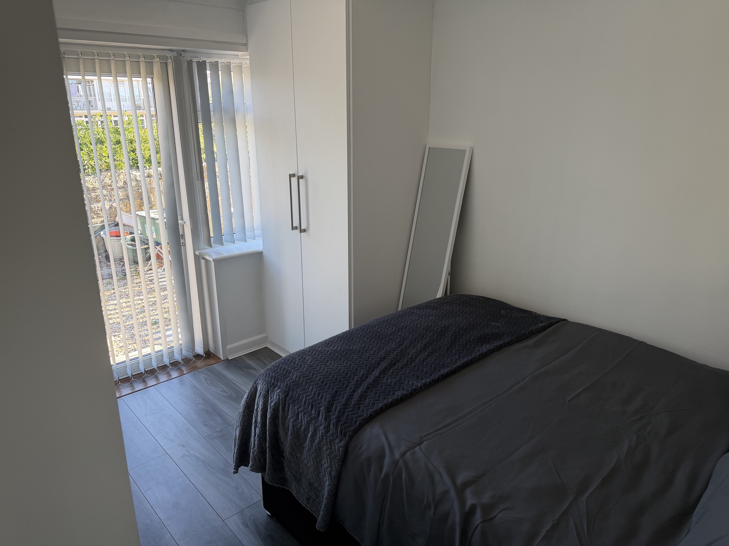 A bedroom with a black bedspread, a wardrobe, a mirror leaning against the wall, and a window with vertical blinds showing a view of a residential area outside.