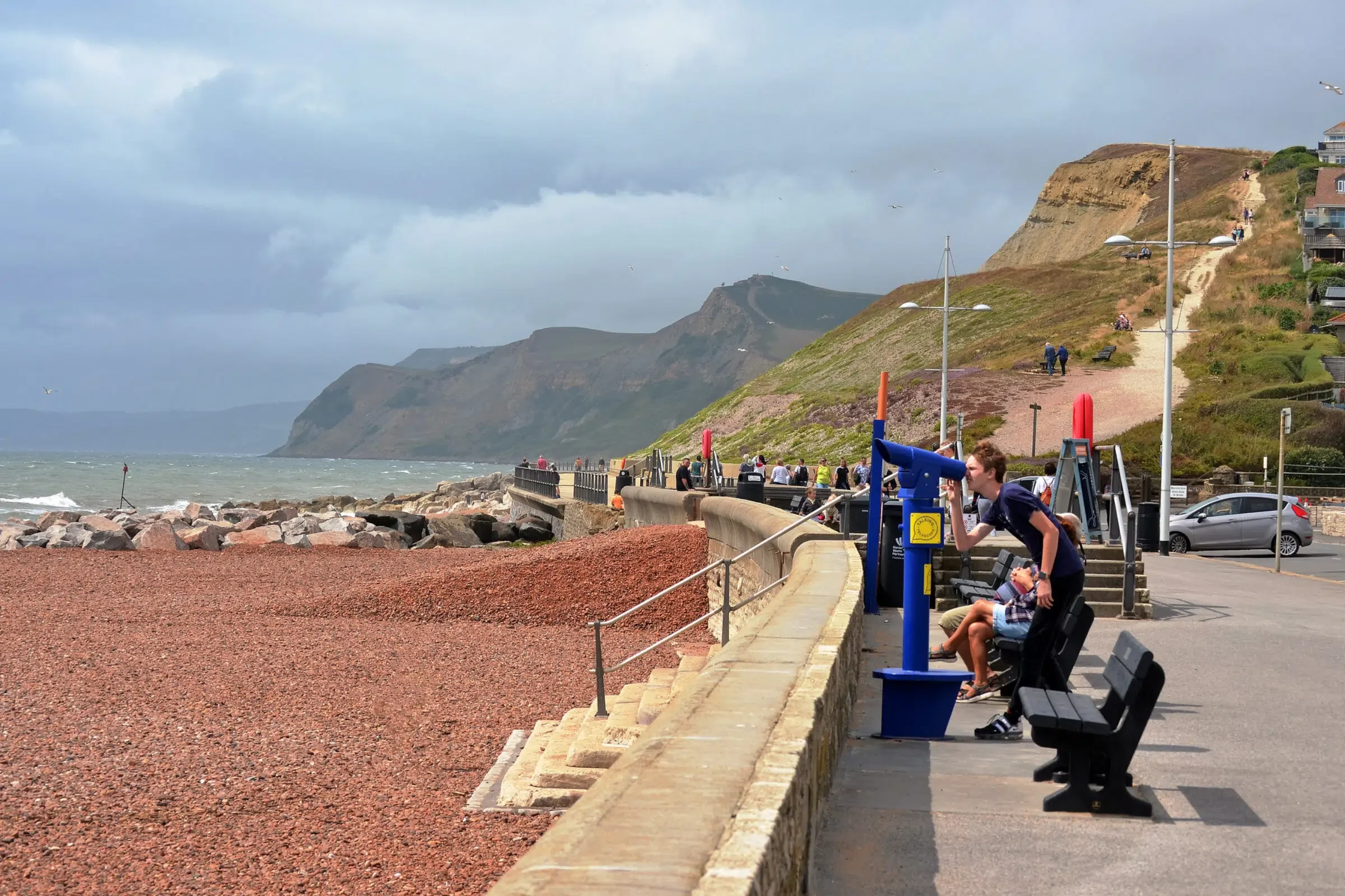 Seaside promenade with sea view, rocky shoreline, people sitting and walking, hillside with stairs and houses, cloudy sky, and streetlights.
