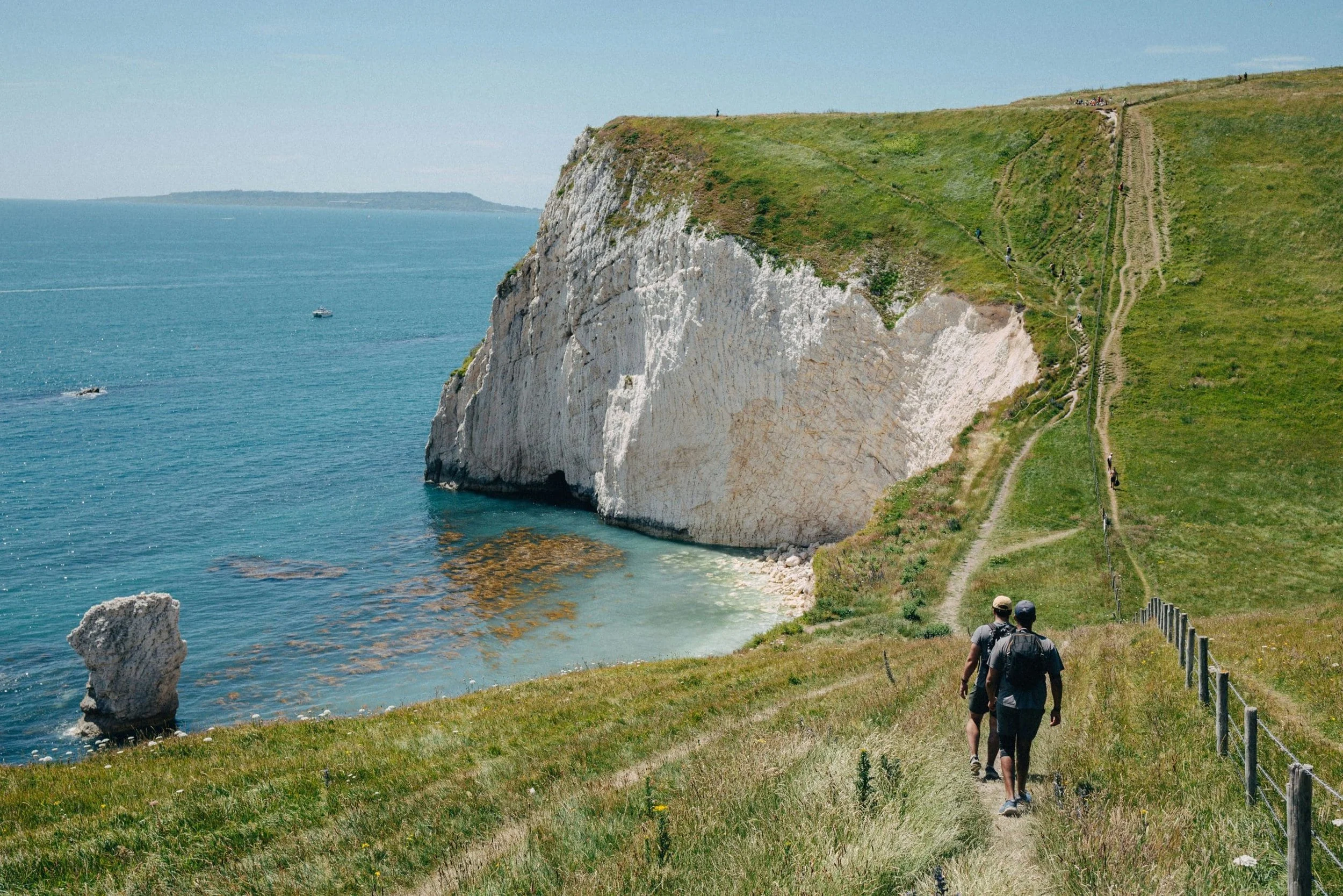 Two hikers walking along a grassy coastal trail with a white chalk cliff face on the right and the ocean in the background on the left.