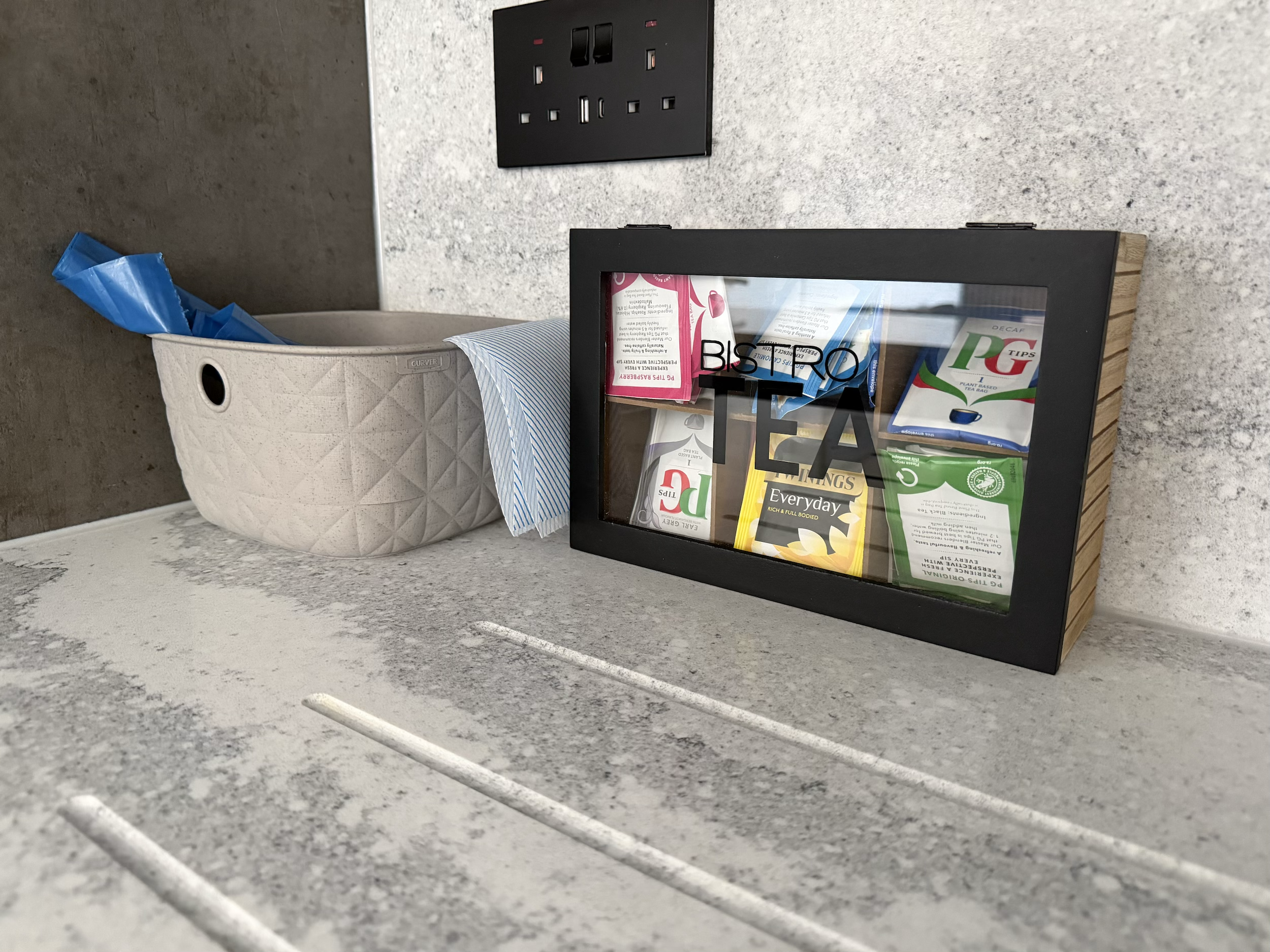 A countertop with a tissue box, a small black frame with various teas, and a wall socket in the background.