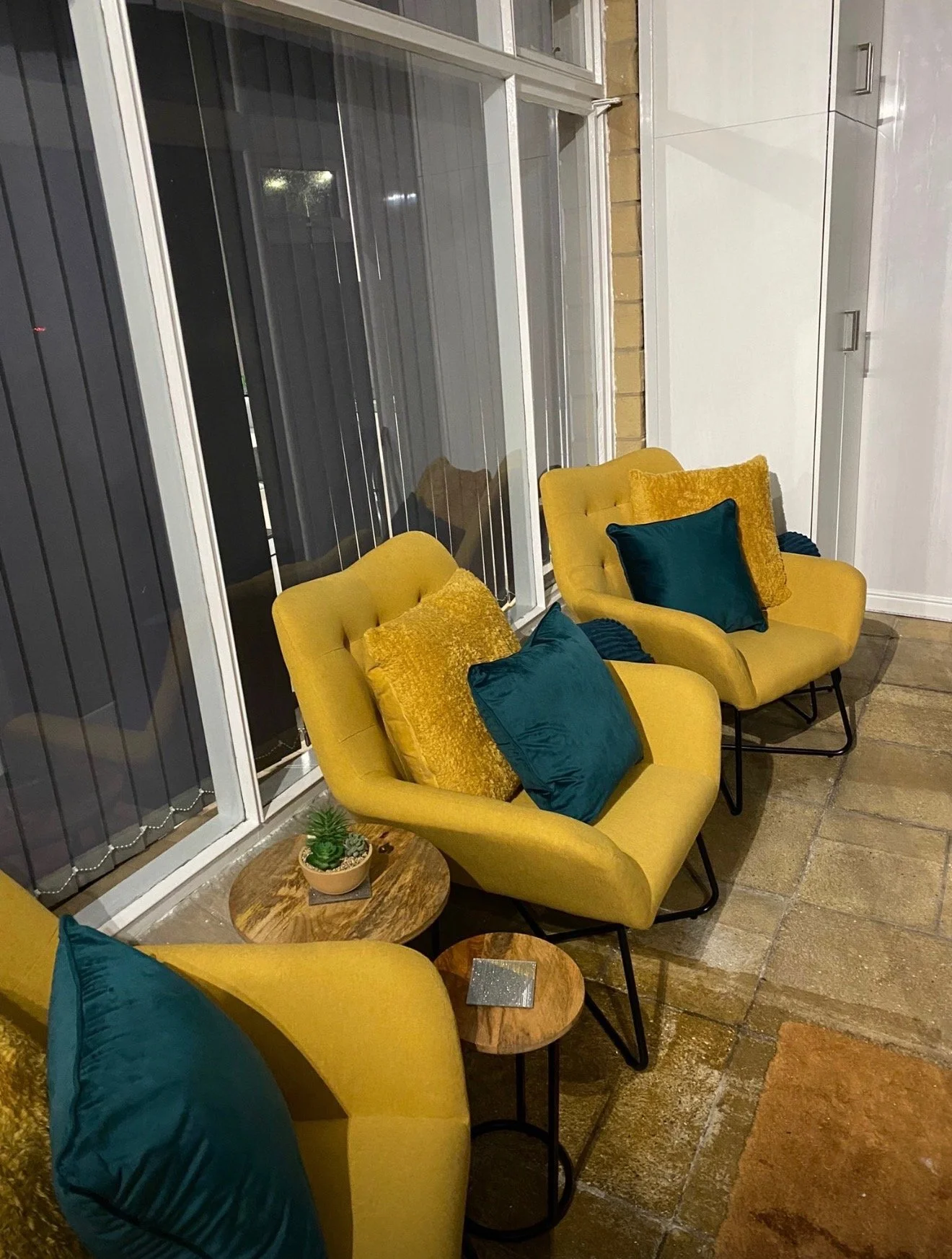 Interior of a seating area with three yellow armchairs with teal and gold pillows, small wooden side tables with potted succulent plants, brick wall, and glass sliding doors with vertical blinds.