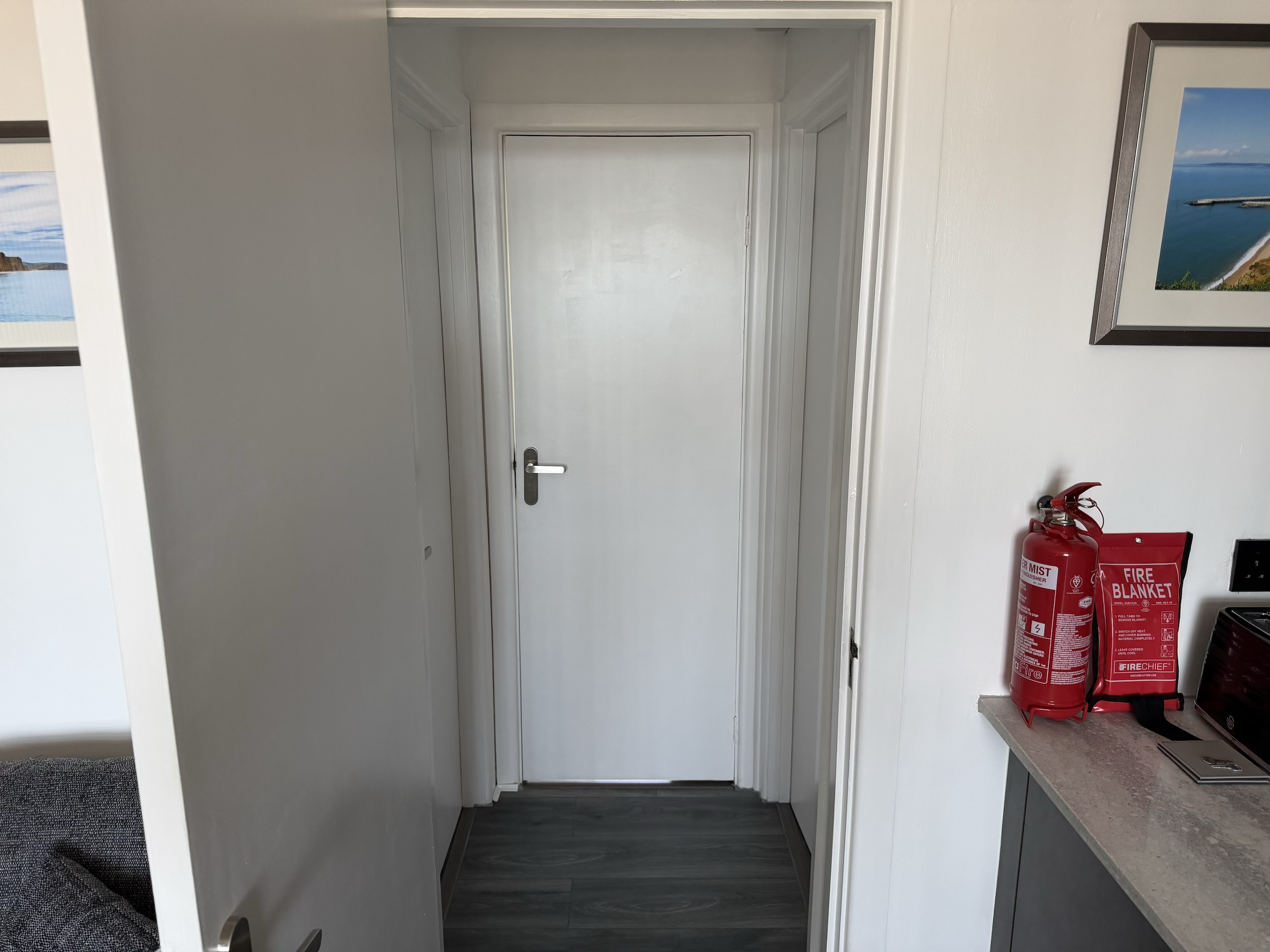 Empty white door with handle in a room painted white, with picture frames on the wall to the right and a fire extinguisher and red fire blanket in the corner near the counter.