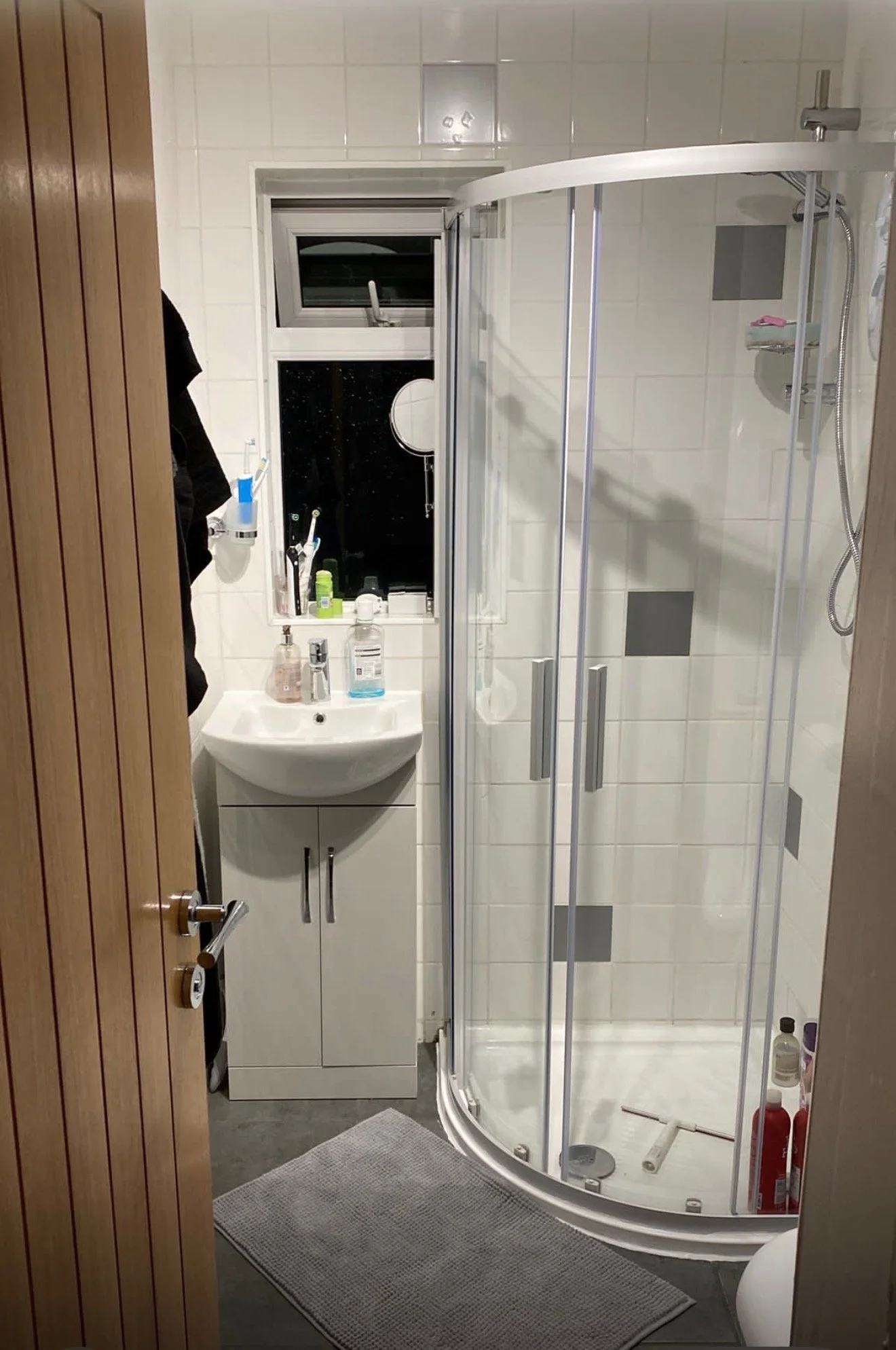 A small bathroom with a shower cabin, a small white sink with a mirror above it, a window above the sink, and various toiletries on the sink and windowsill. The bathroom has grey tiles and a wooden door with a silver handle.