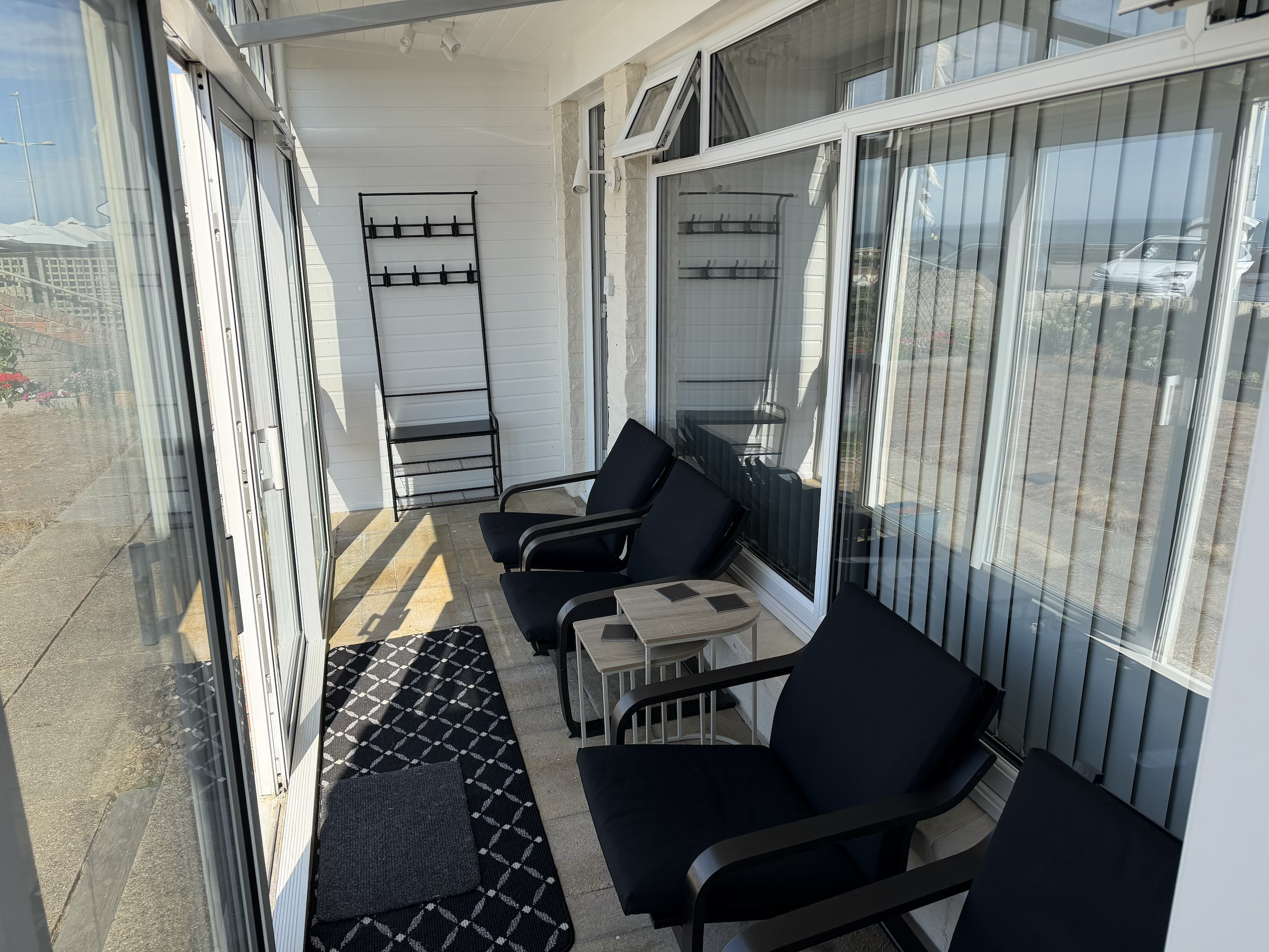 Sunlit enclosed porch with black chairs, a small table, and a black rack on white siding wall, with large glass windows and door reflecting parked cars outside.