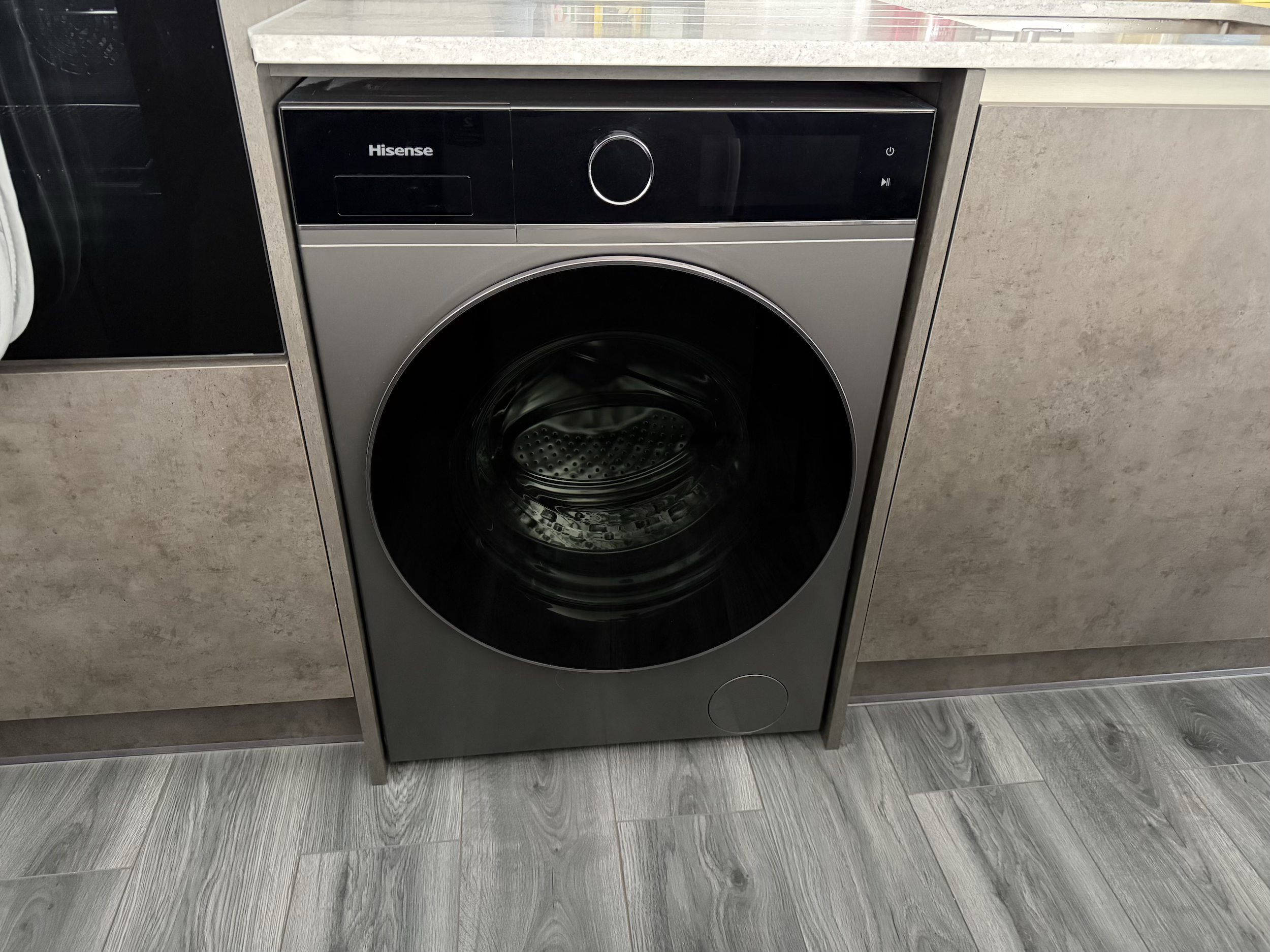 A modern front-loading Hisense washing machine built into a beige kitchen or laundry room cabinet, with a digital control panel and a round door, on gray wood-look flooring.