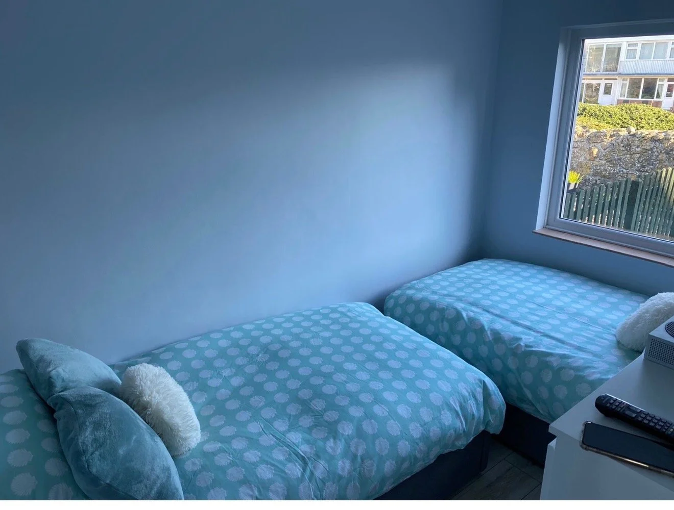 A bedroom with two twin beds featuring blue and white polka dot bedding, pillows, and stuffed animals. There is a large window showing an outdoor scene with houses and greenery.