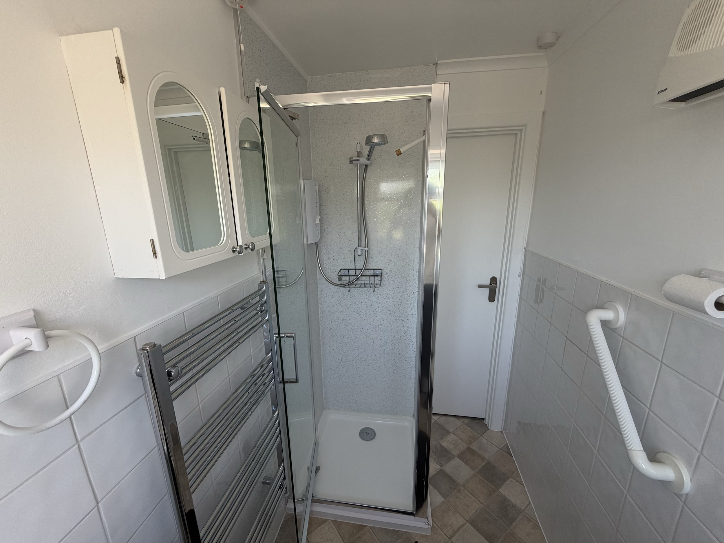 Bathroom with a shower area and glass door, white cabinets above the sink, towel rack, and beige tiled floor.