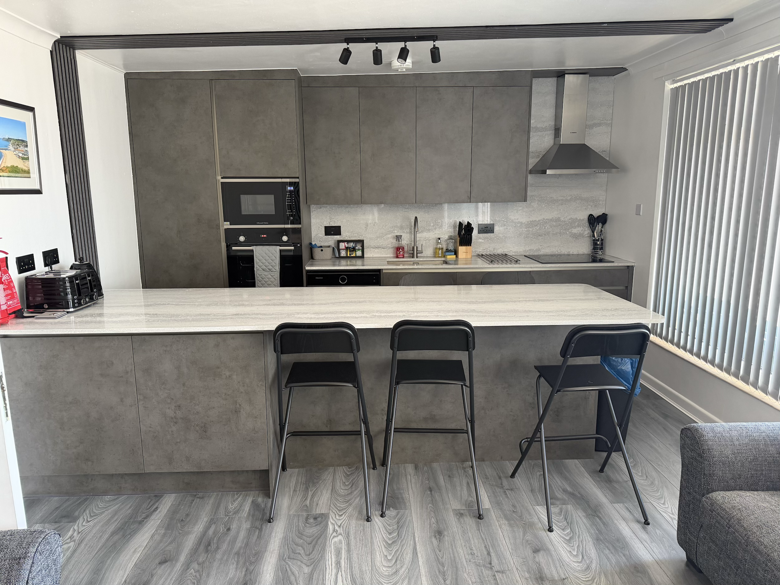 Modern kitchen with grey cabinets, countertop with various kitchen items, and a stainless steel range hood. There are three black chairs in front of the counter, and a window with vertical blinds on the right side.